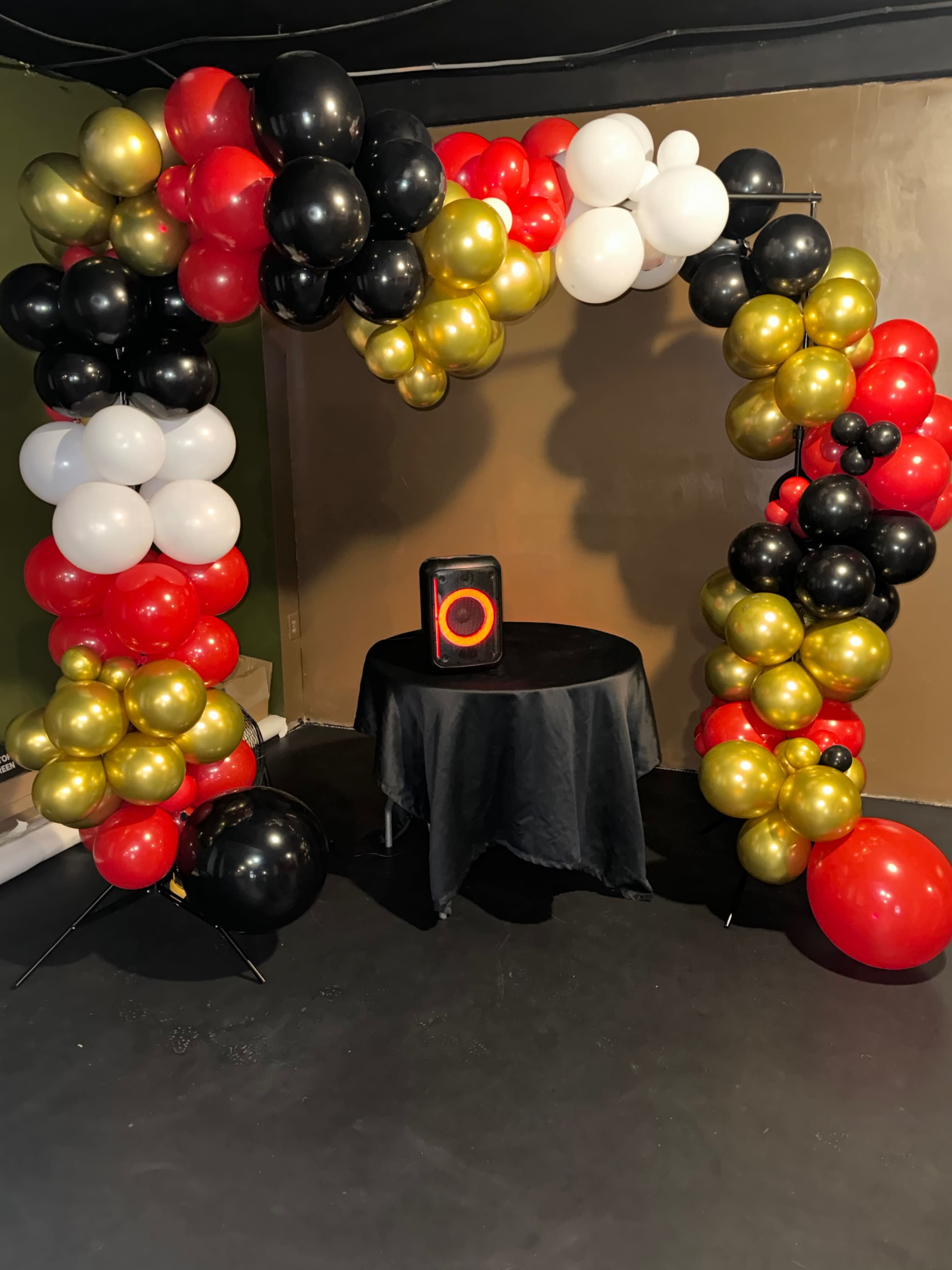 The image shows a decorative arch made of black, white, red, and gold balloons, positioned above a black table with a speaker resting on it.