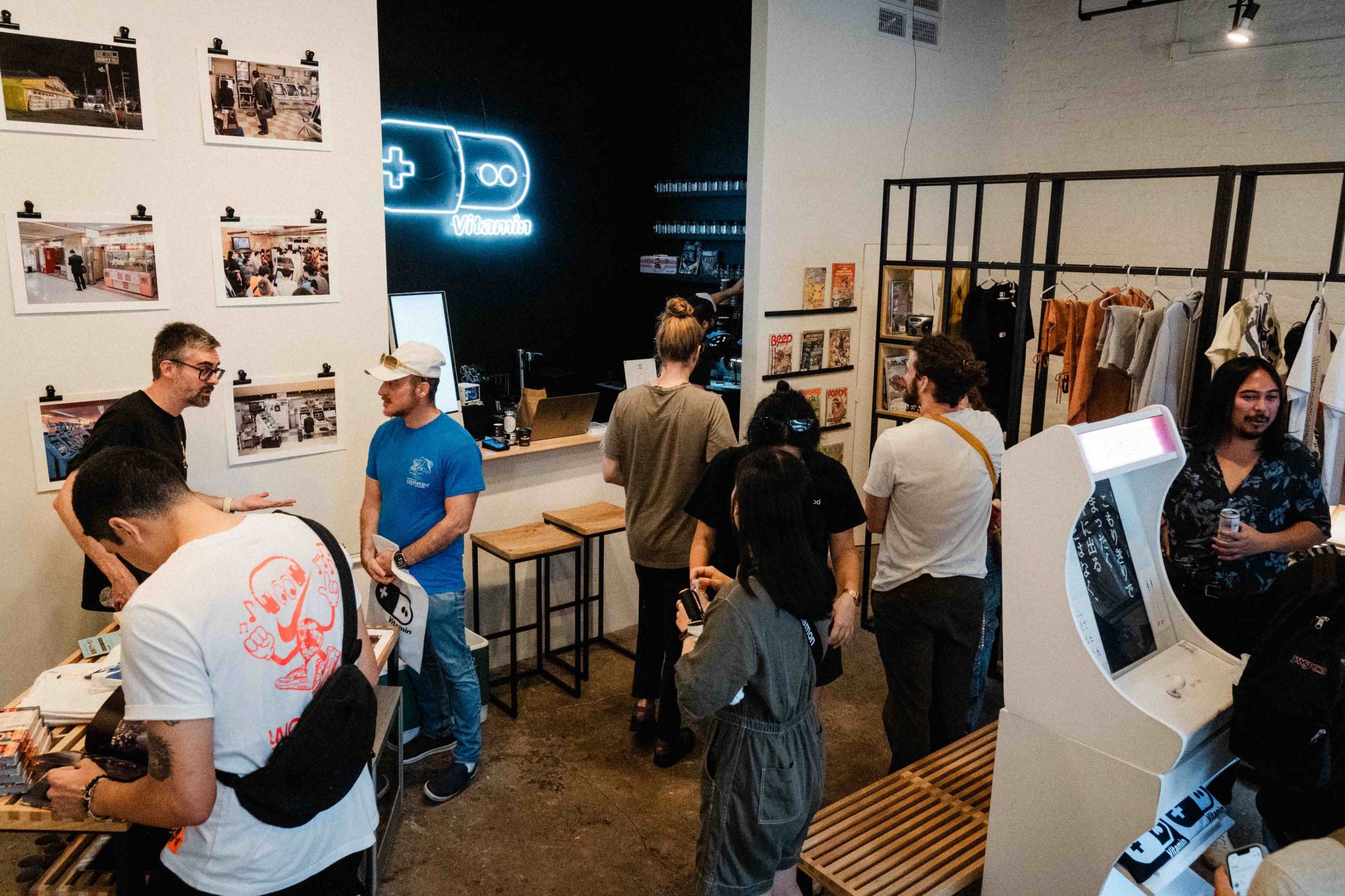 A group of people is gathered inside a retail space featuring a bar area, merchandise displays, and a retro arcade machine.