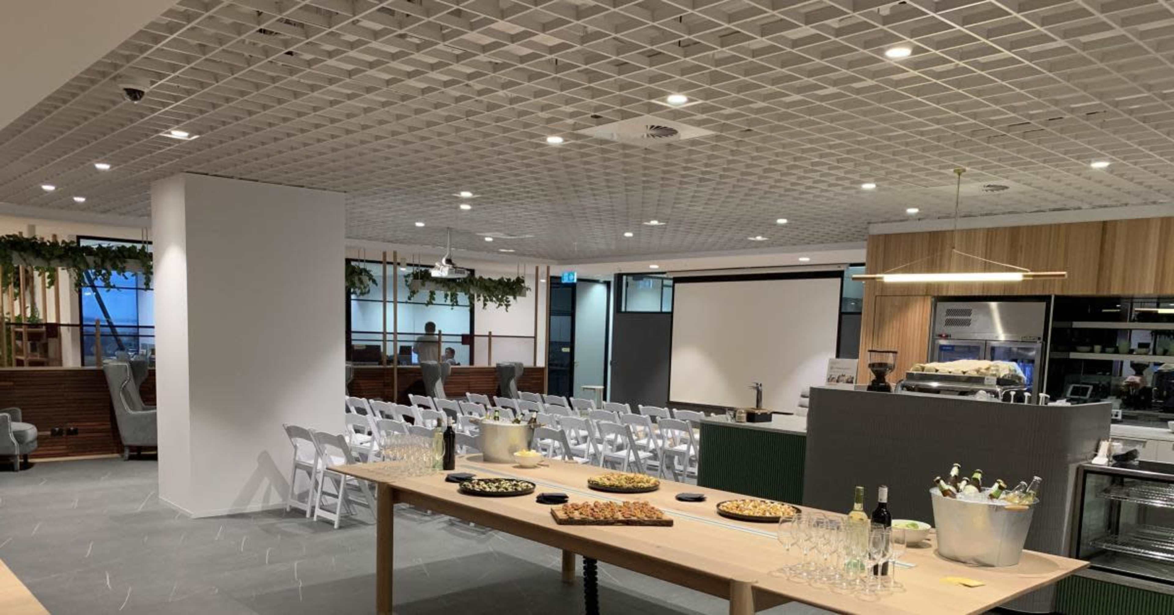 The image shows a modern event space with a long table displaying food and drinks, surrounded by white chairs and a large screen against a backdrop of contemporary décor.