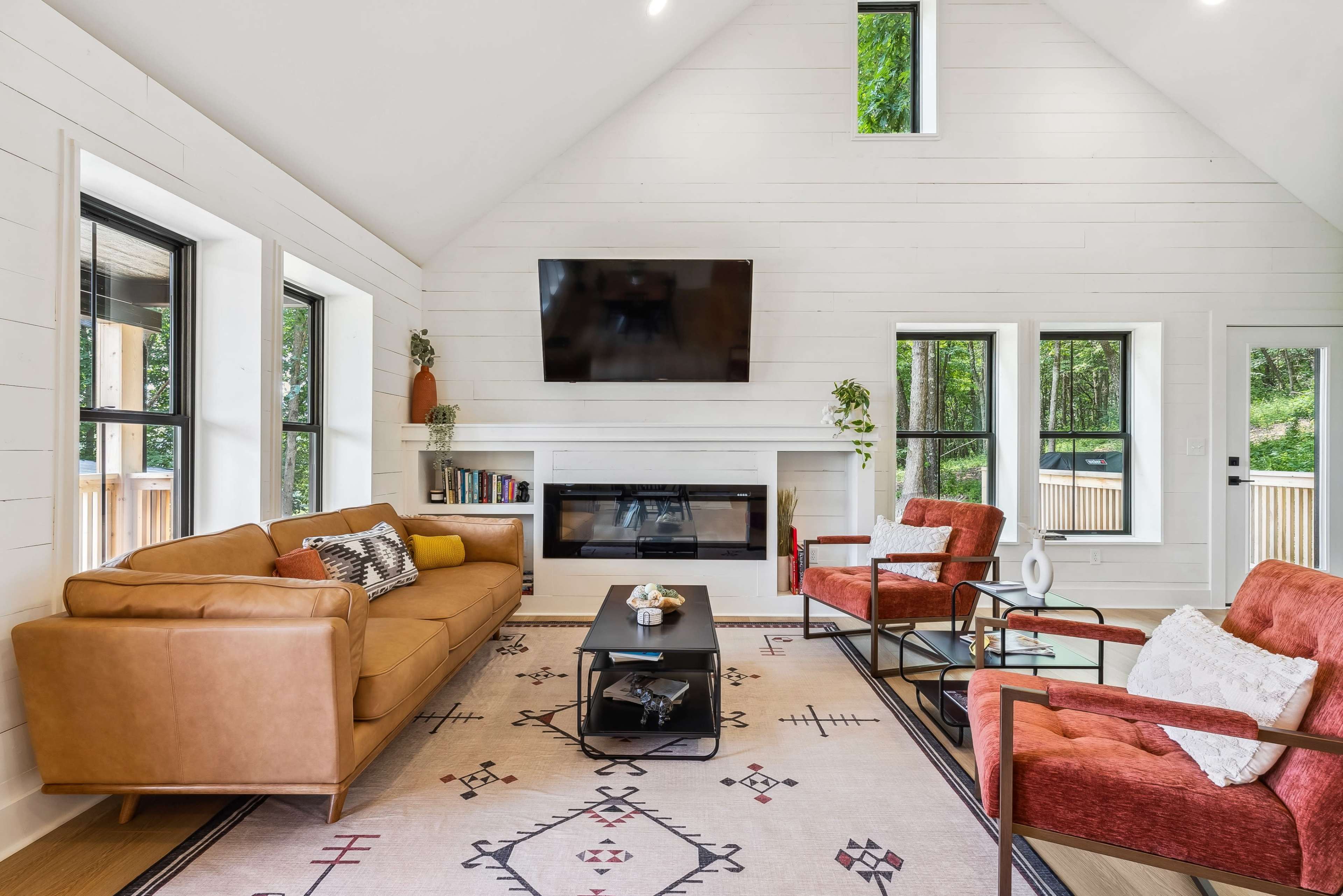 The living room features a brown sofa, two red armchairs, a black coffee table, and a mounted television above a modern fireplace, with large windows allowing natural light to enter.