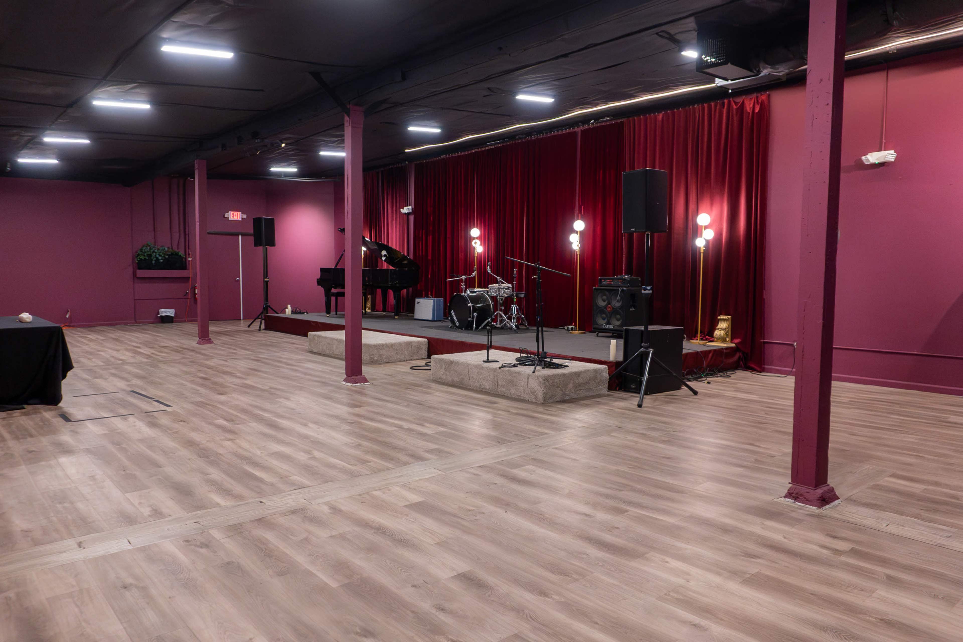 The image shows a music performance stage with a grand piano, drum set, and sound equipment, set against a backdrop of red curtains in a room with wooden flooring.