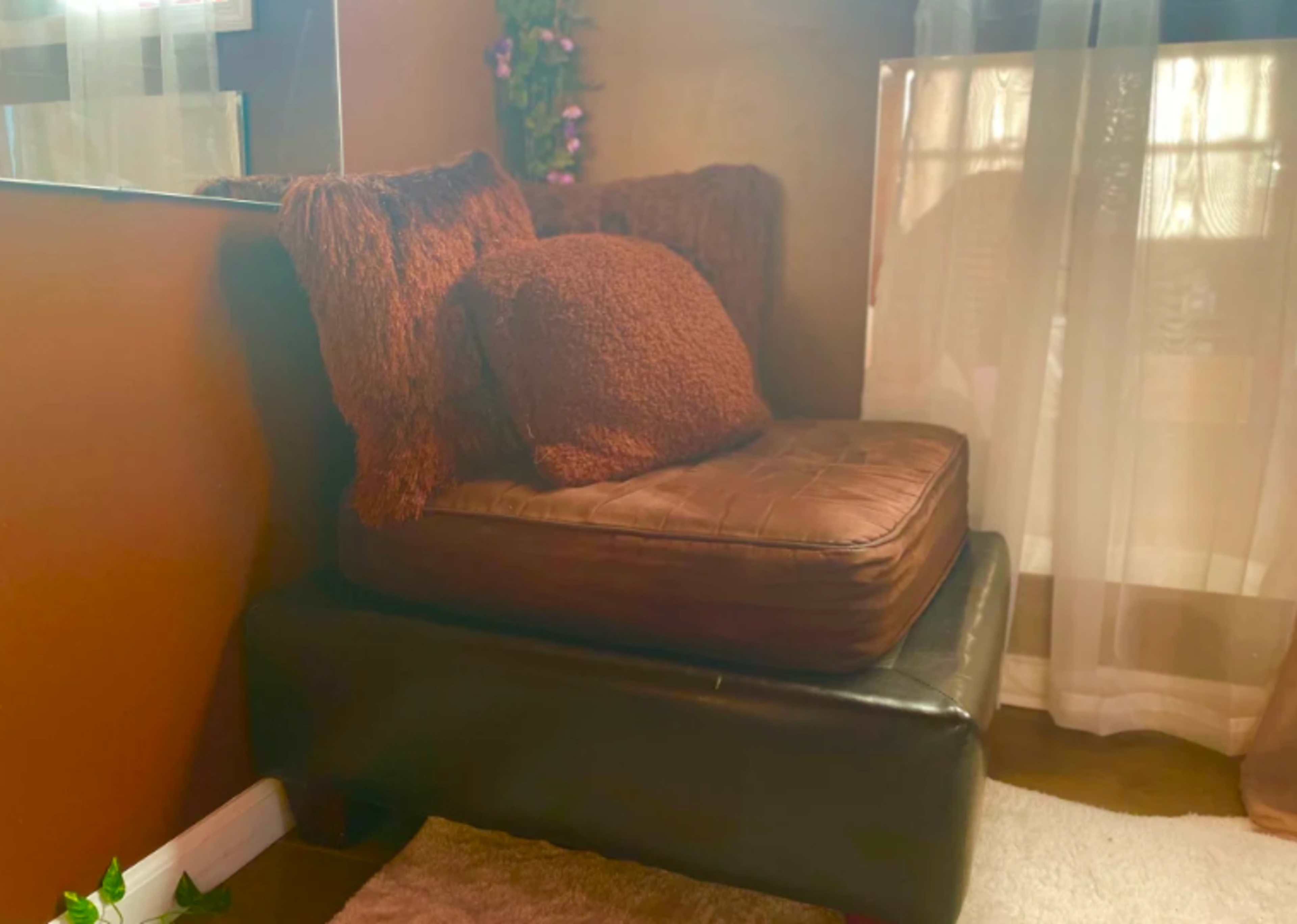A cozy seating area with a brown cushioned chair stacked on a black ottoman, positioned near a window with sheer curtains.