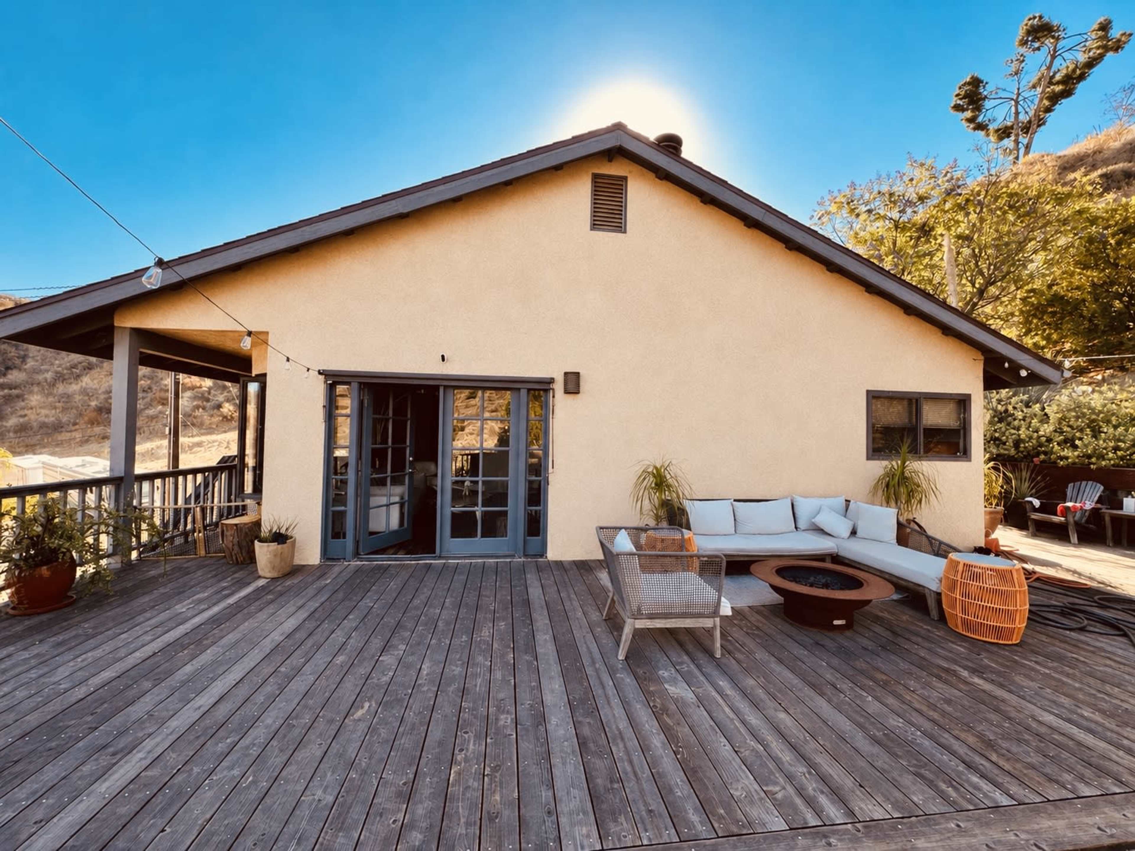 The image shows a cozy wooden deck attached to a light-colored house, featuring outdoor seating and a fire pit surrounded by potted plants.