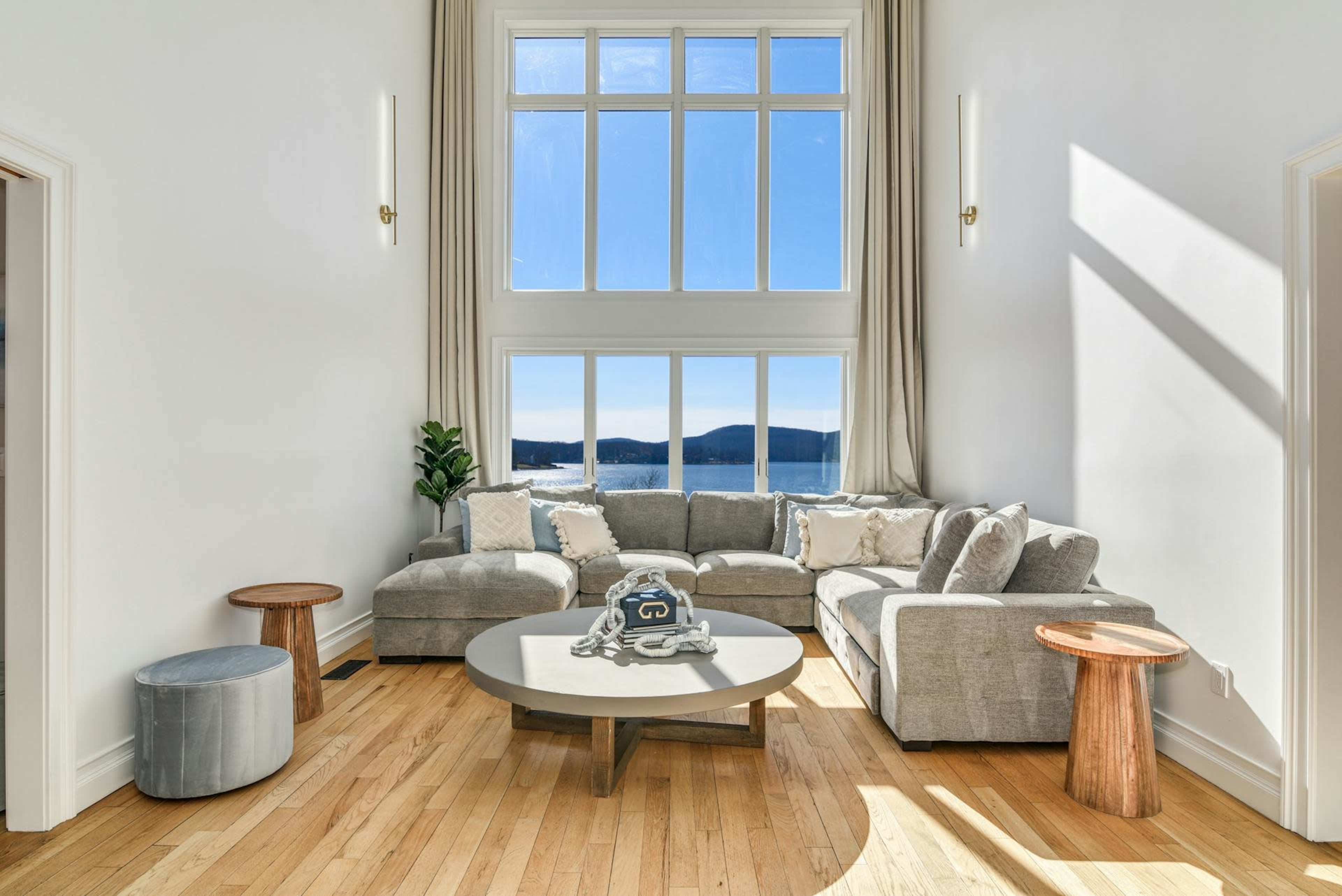 The room features a large, light-filled corner with a sectional sofa, a circular coffee table, and two wooden side tables, overlooking a scenic view through tall windows.