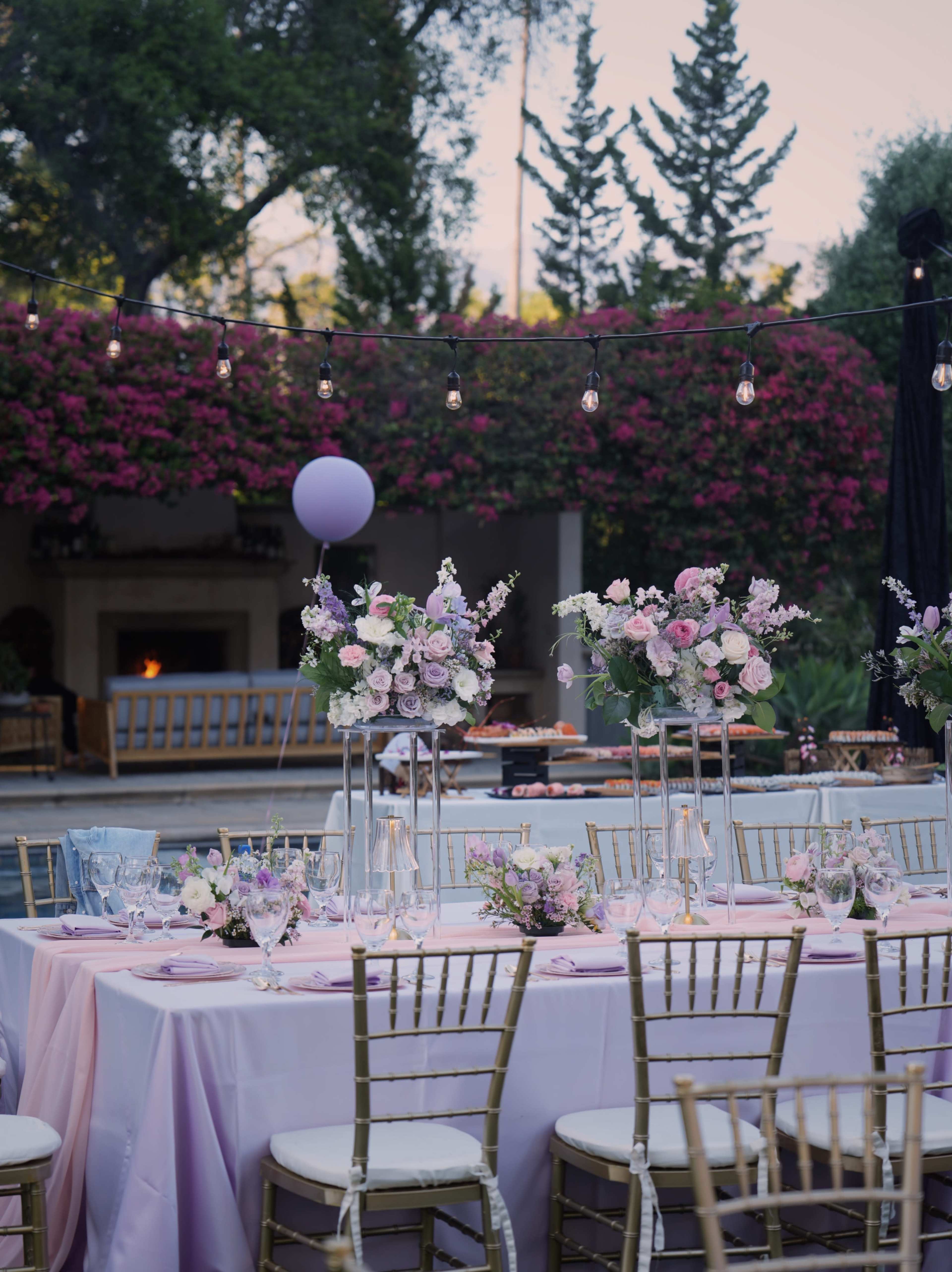 A beautifully set outdoor dining table is adorned with elegant floral centerpieces and surrounded by gold chairs, with string lights and blooming greenery in the background.