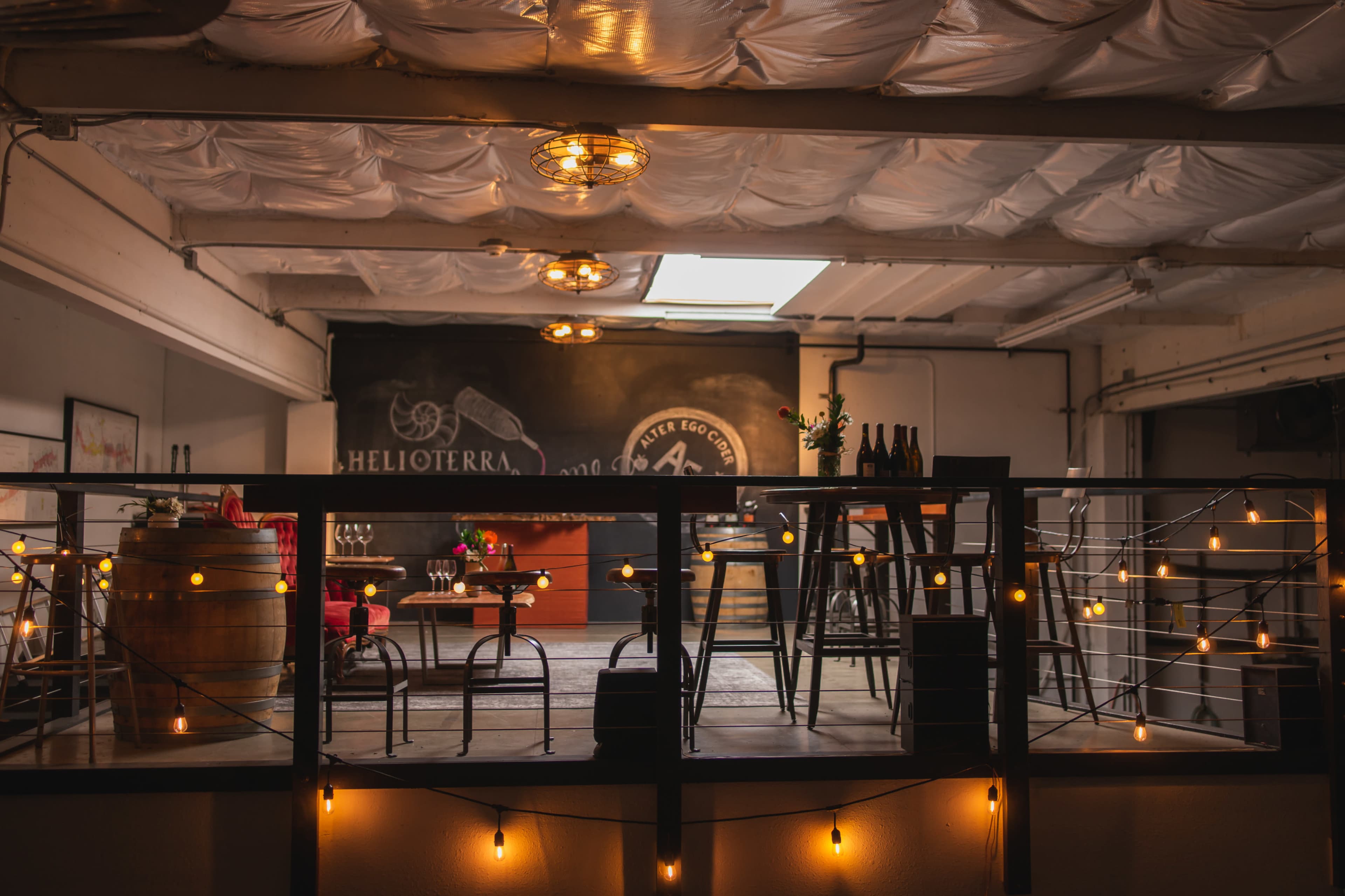 A cozy upper level of a bar or wine tasting room featuring wooden furniture, string lights, and a chalkboard background.