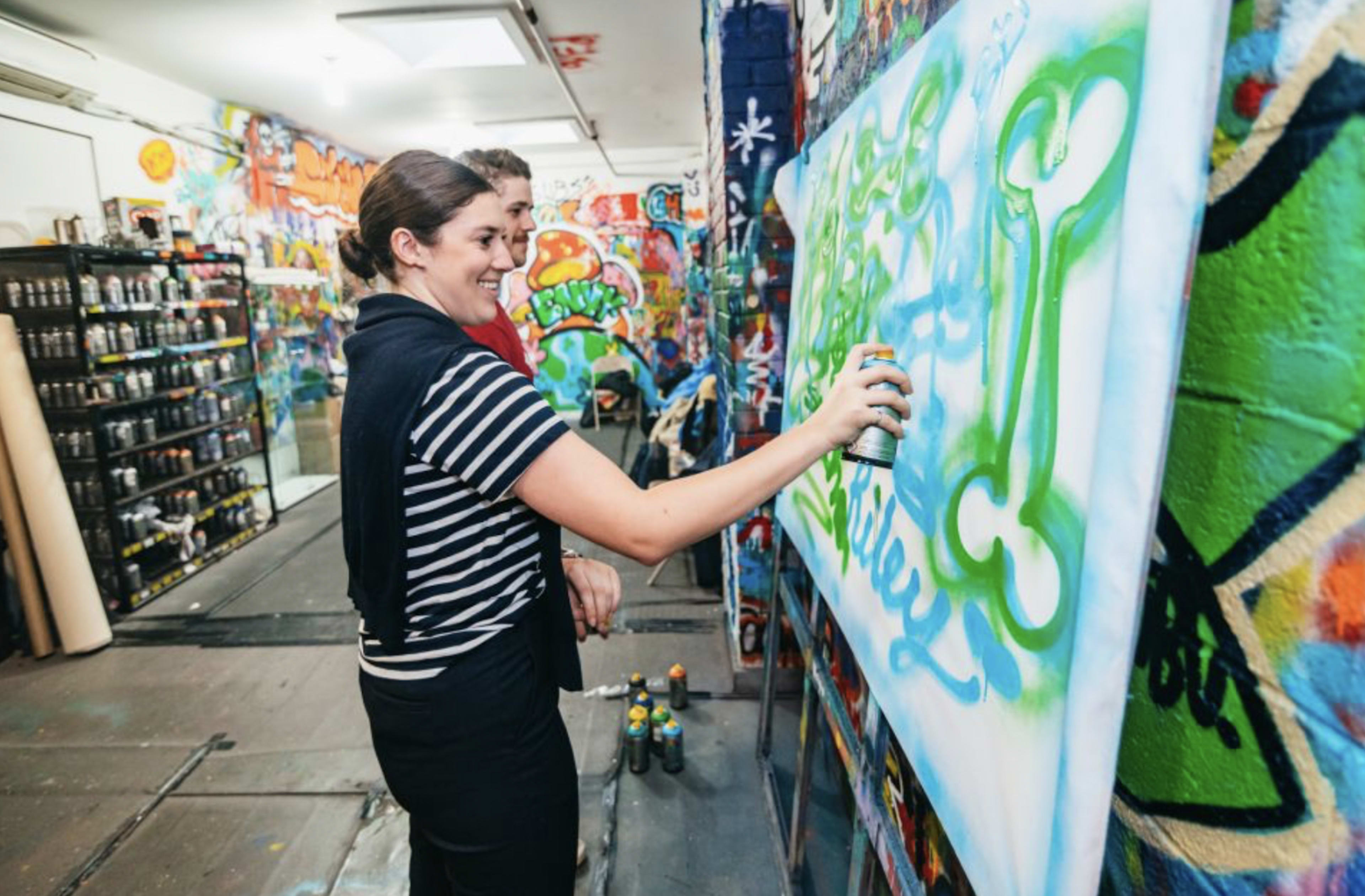 A woman sprays paint onto a canvas while a man observes in a graffiti art studio filled with colorful murals.