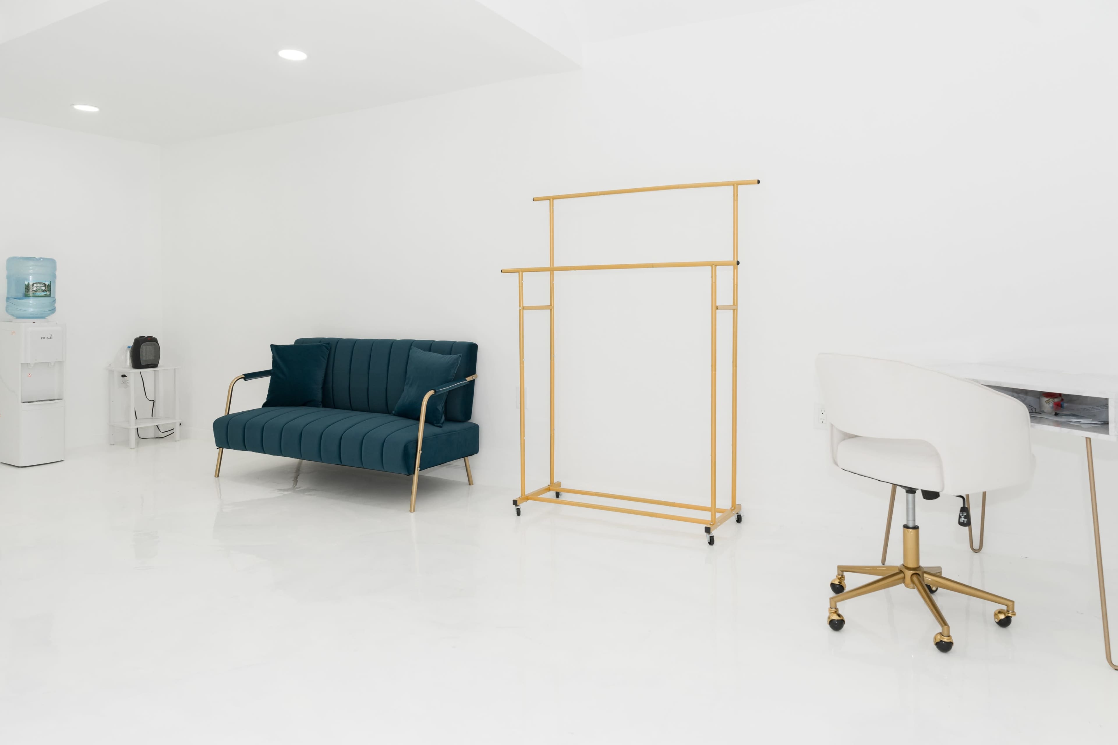 A minimalist room with a teal sofa, a gold clothing rack, a white desk chair, and a water cooler.