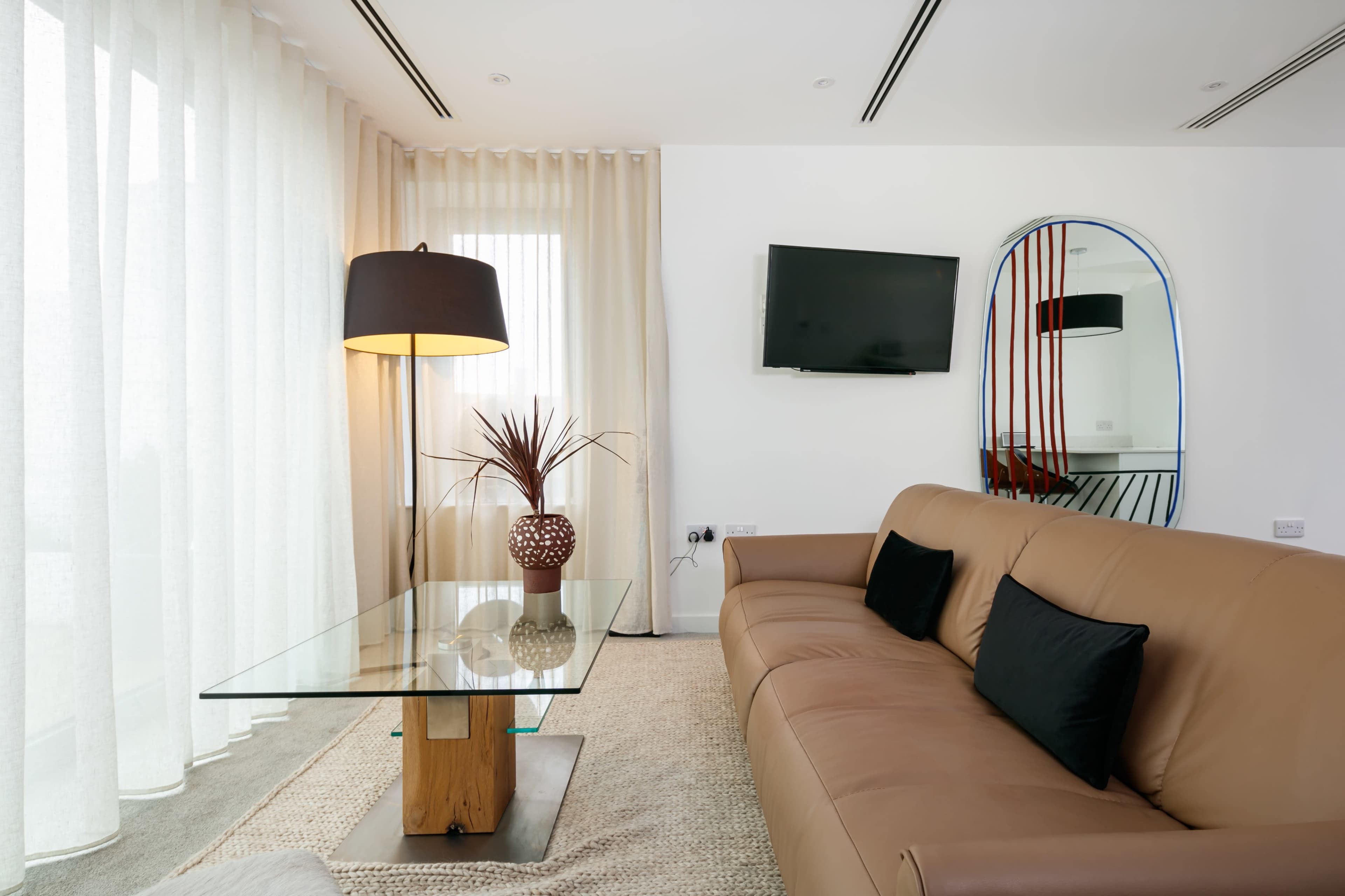 A modern living room with a brown sofa, glass coffee table, a decorative plant in a vase, a floor lamp, a wall-mounted television, and sheer curtains.