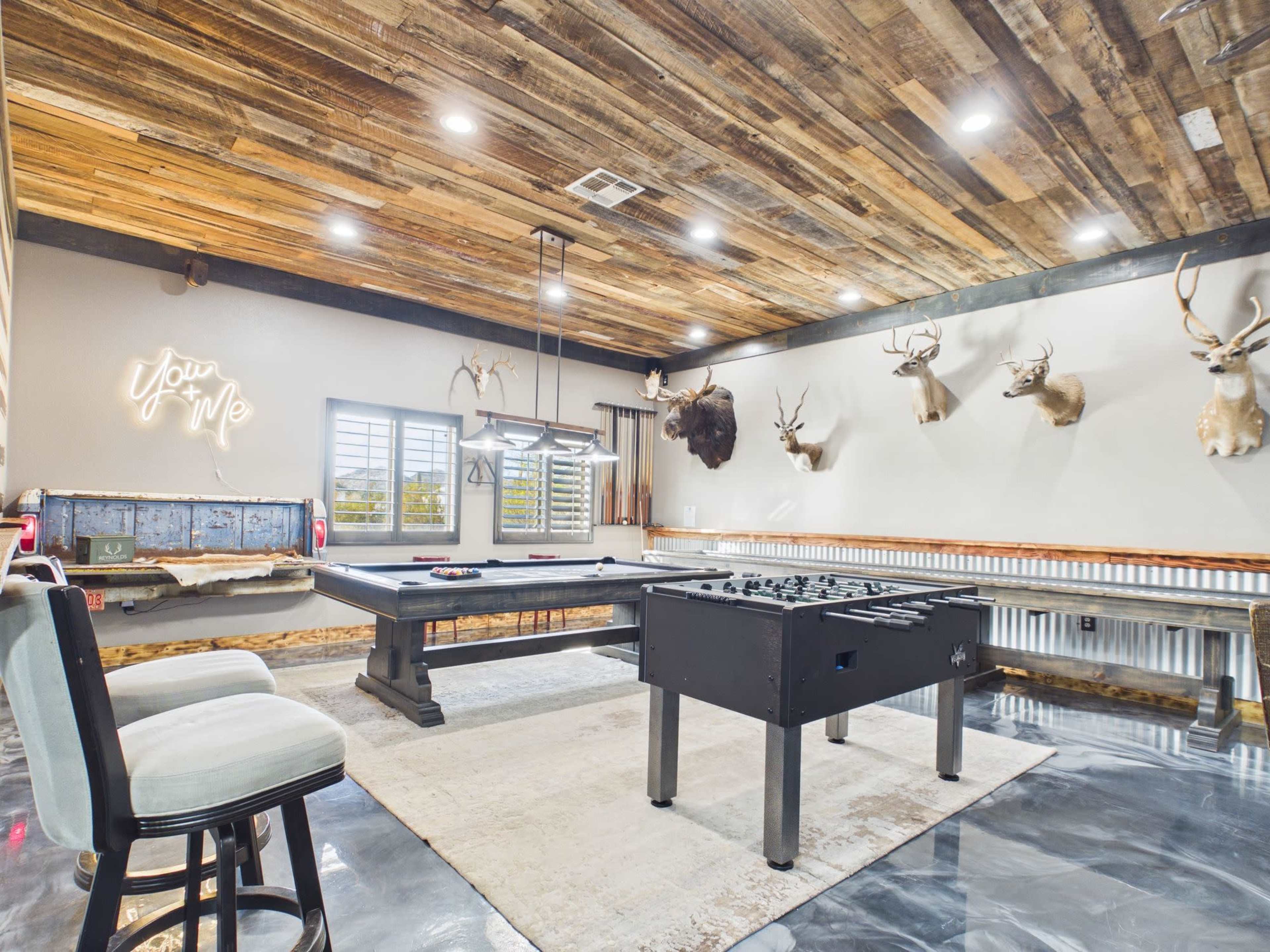 The image shows a game room featuring a pool table, a foosball table, and several mounted deer heads on the walls, with wooden ceiling beams and a rustic decor style.