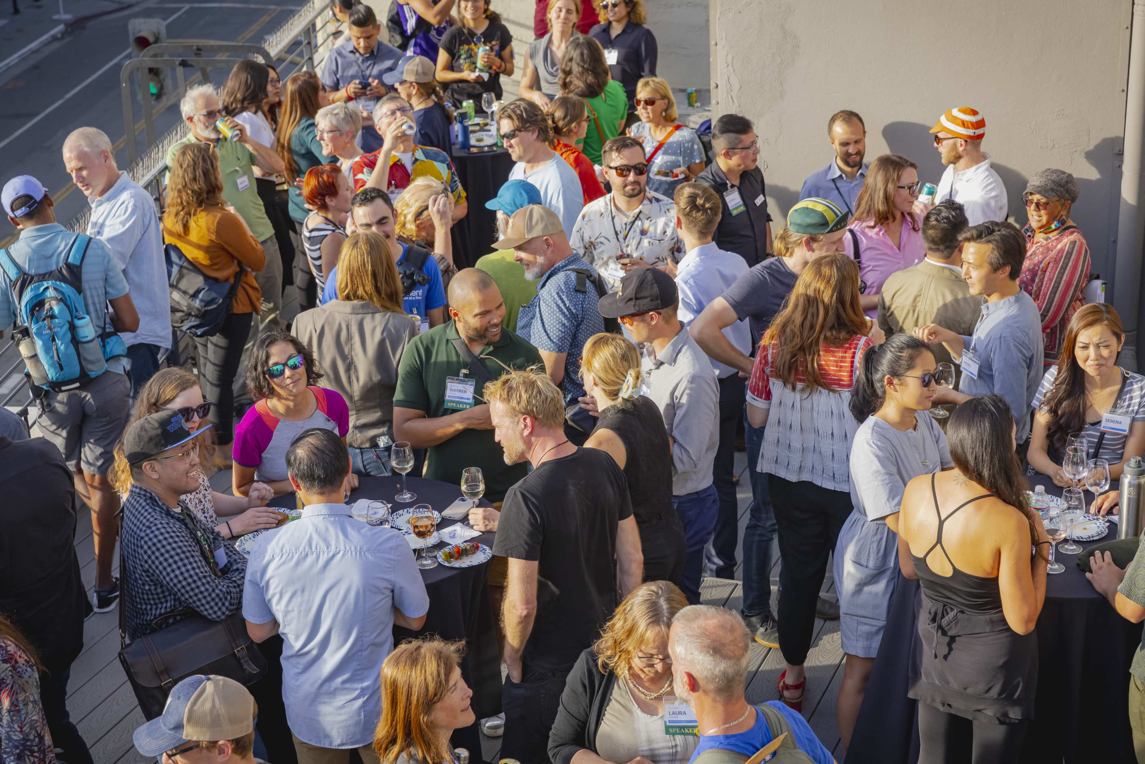 A large group of people mingles at an outdoor event, standing around high tables filled with drinks and snacks.