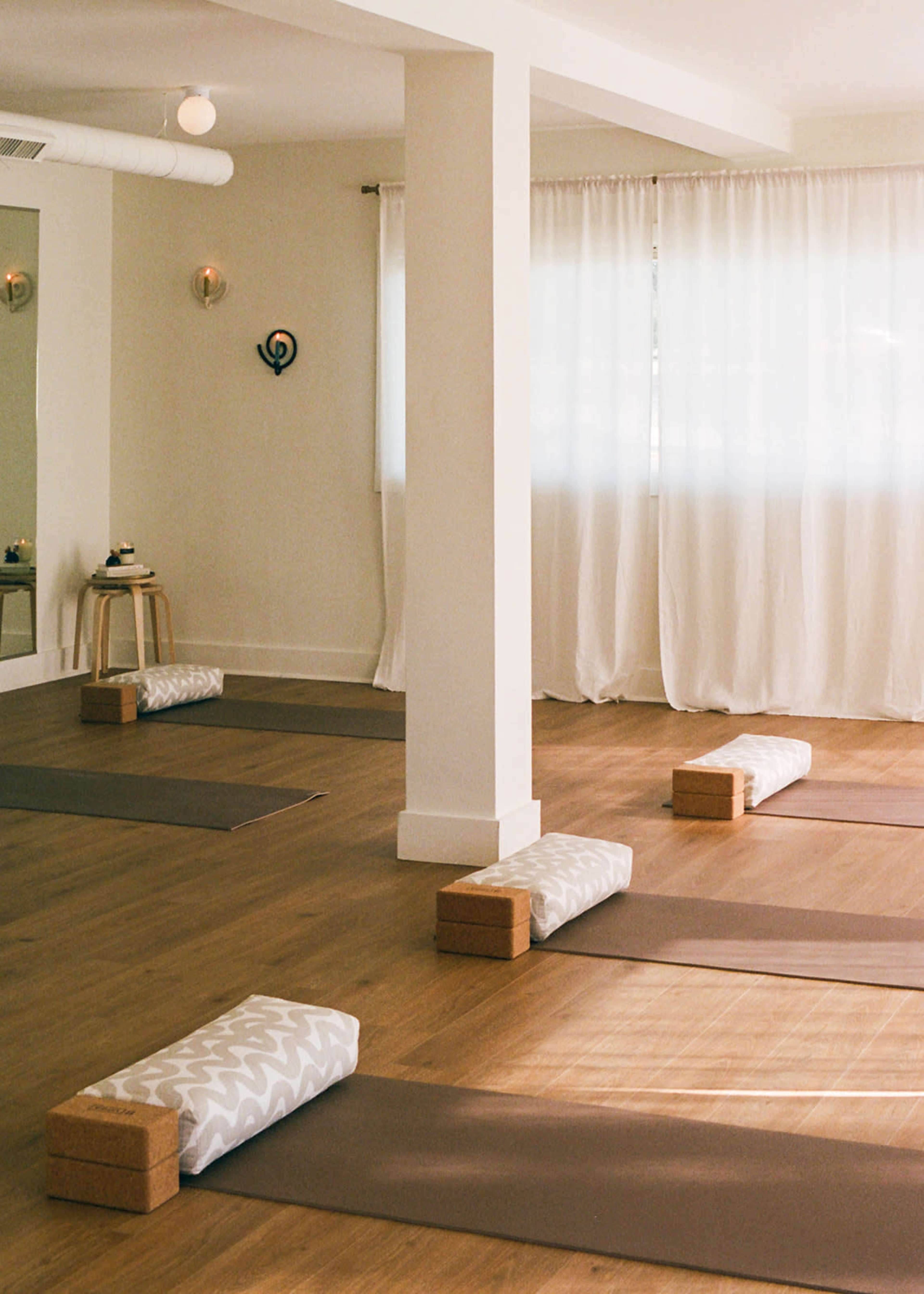 The image shows a serene yoga studio with several yoga mats and props arranged neatly on a wooden floor, illuminated by natural light through sheer white curtains.