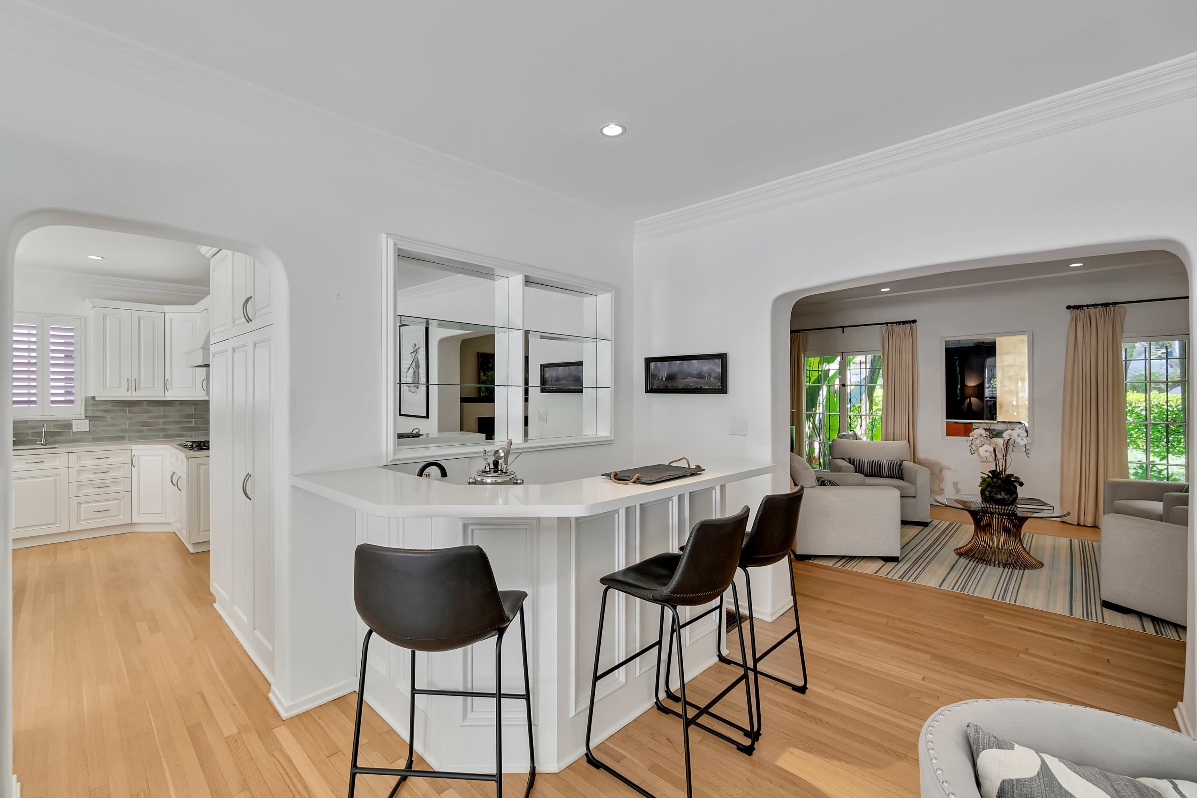 The image shows a modern, open-concept living space with a kitchen bar area and a view of a sunlit, furnished living room.