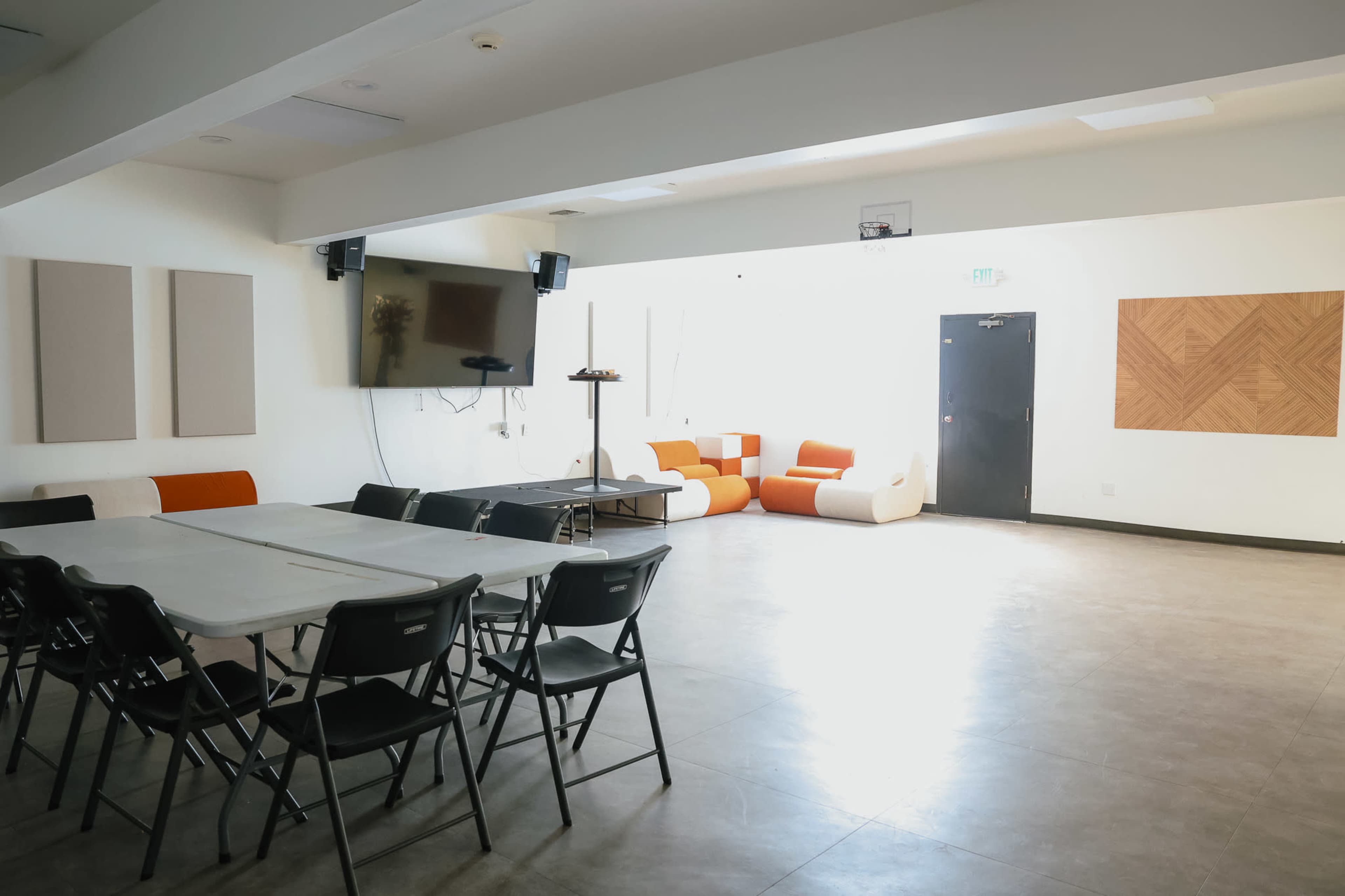 The image shows a spacious room with a long table surrounded by black chairs, a large screen on the wall, and a seating area with orange and white couches.