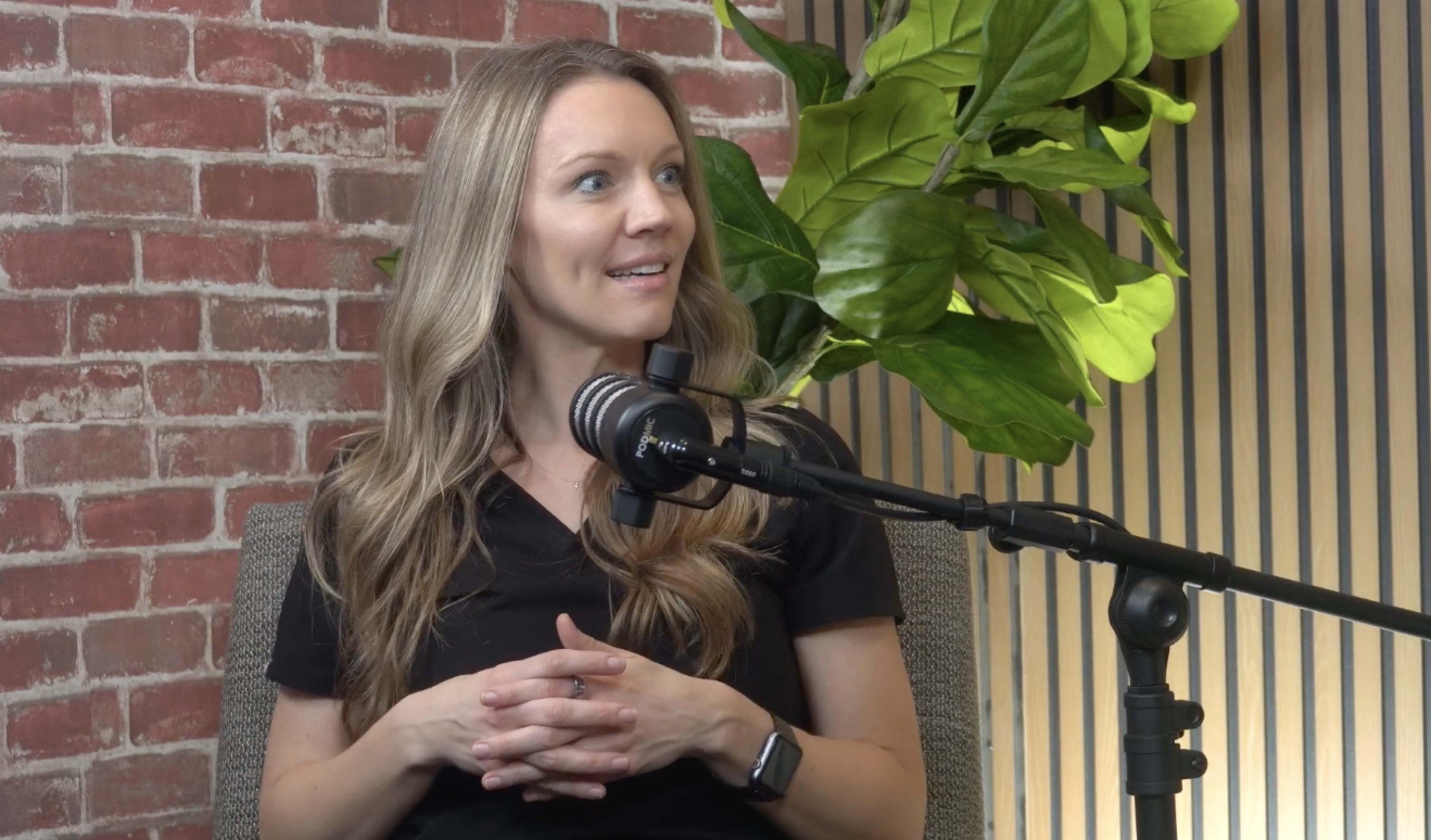 A woman with long, wavy hair sits in front of a microphone, surrounded by a brick wall and green foliage.