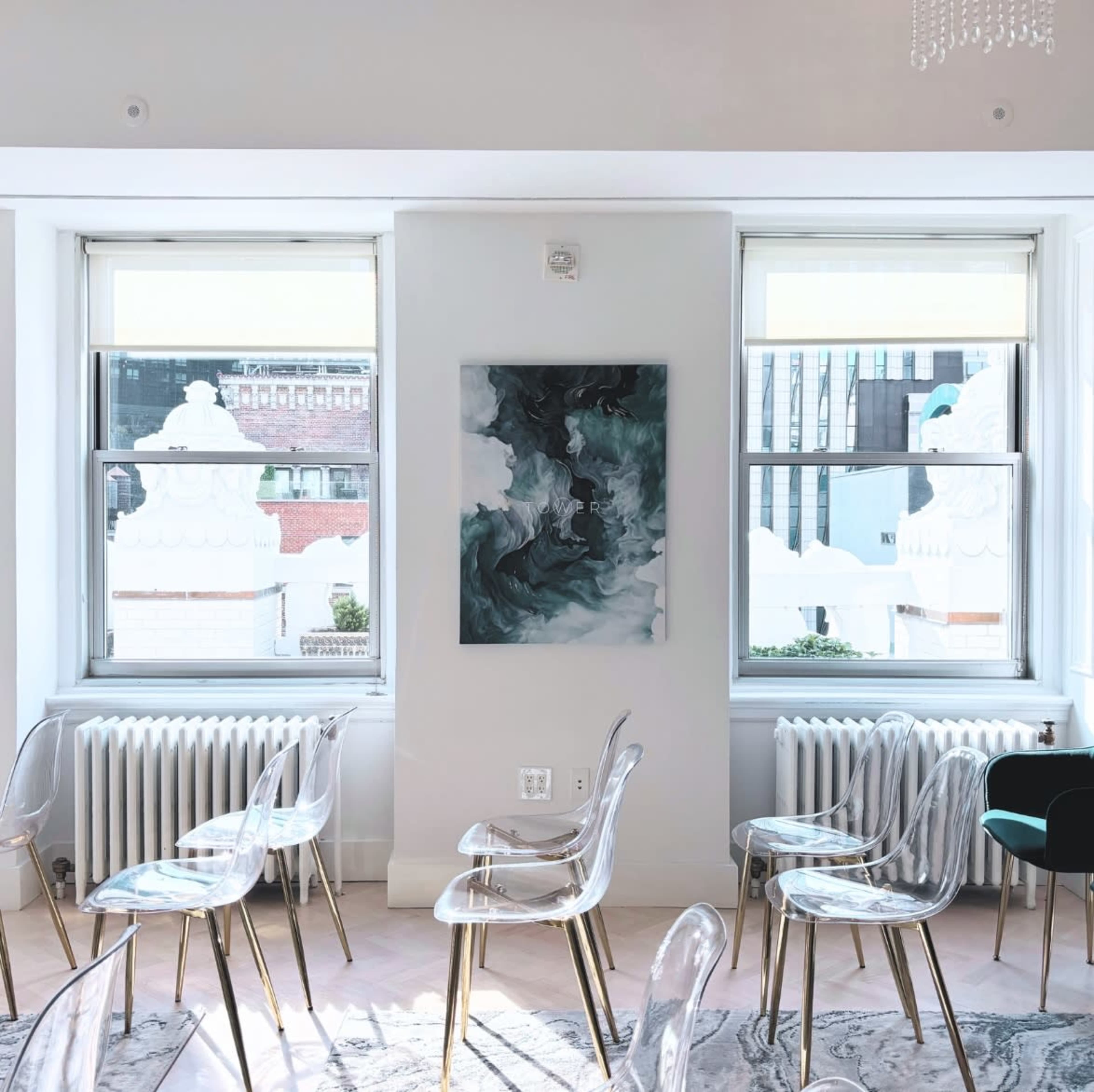 A bright room features clear plastic chairs arranged in rows, with large windows displaying a cityscape and a painting on the wall.