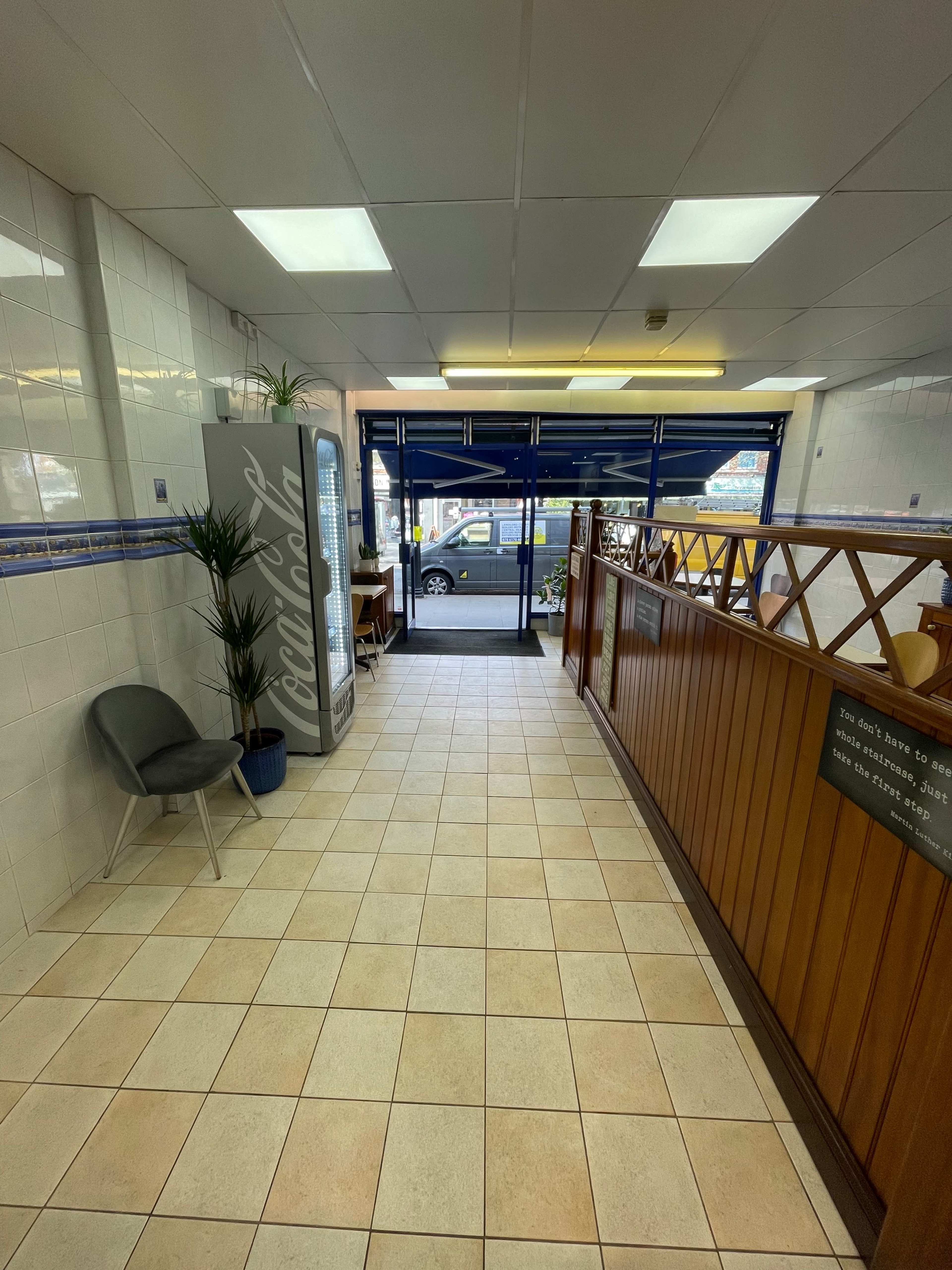 A tiled hallway in a restaurant with a vending machine, a plant, and wooden railing leading to an entrance.