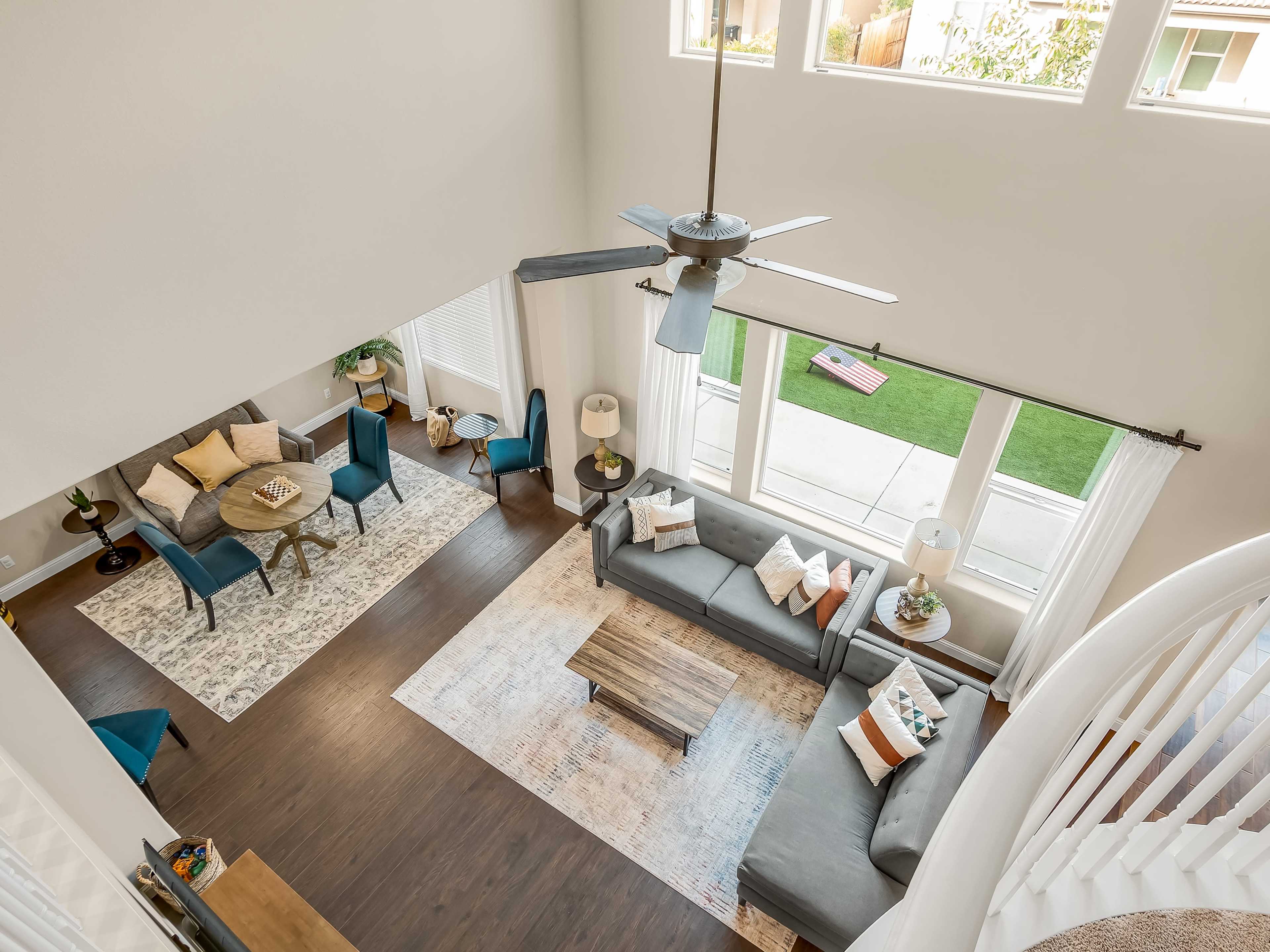 A spacious living area with a high ceiling, featuring a gray sofa, a wooden coffee table, a dining table with blue chairs, and large windows allowing natural light.