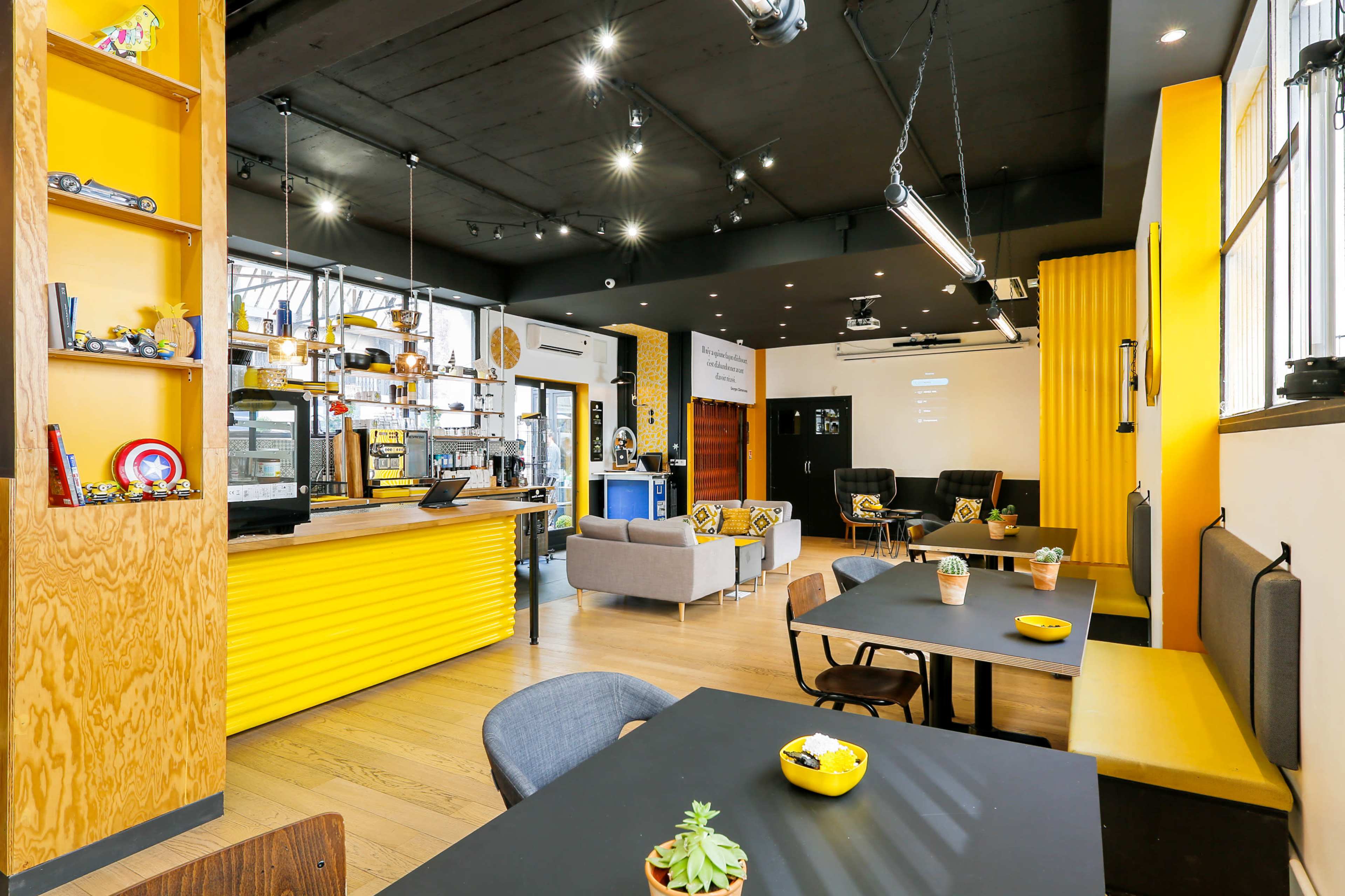 The image shows a modern café with yellow and wood accents, featuring seating areas, a coffee counter, and decorative shelves.