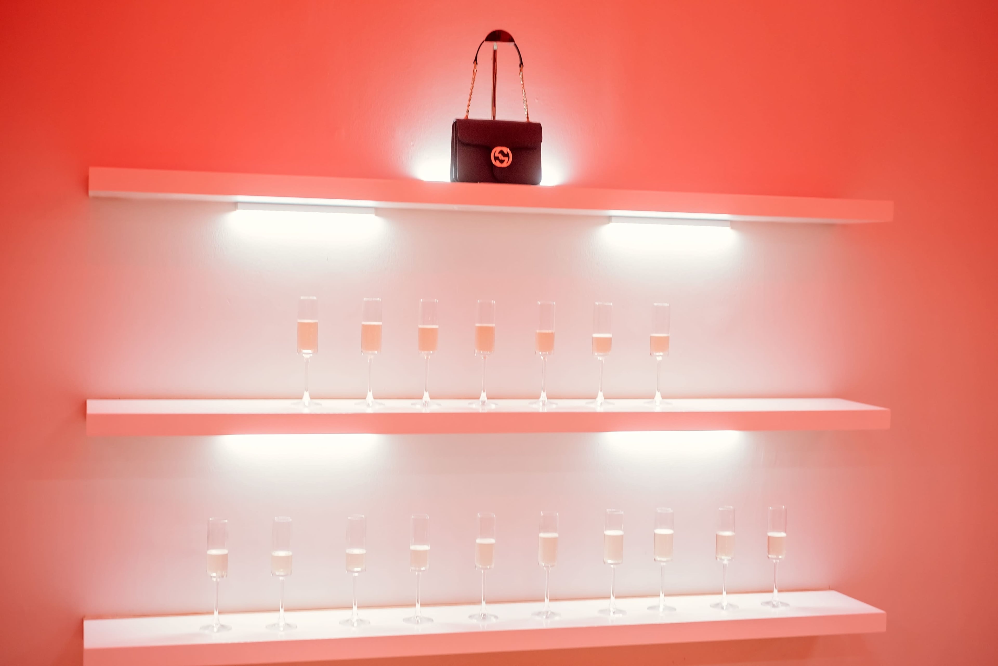 A shelf with a row of champagne flutes, each containing a drink, with a small black handbag displayed above them against a bright red wall.