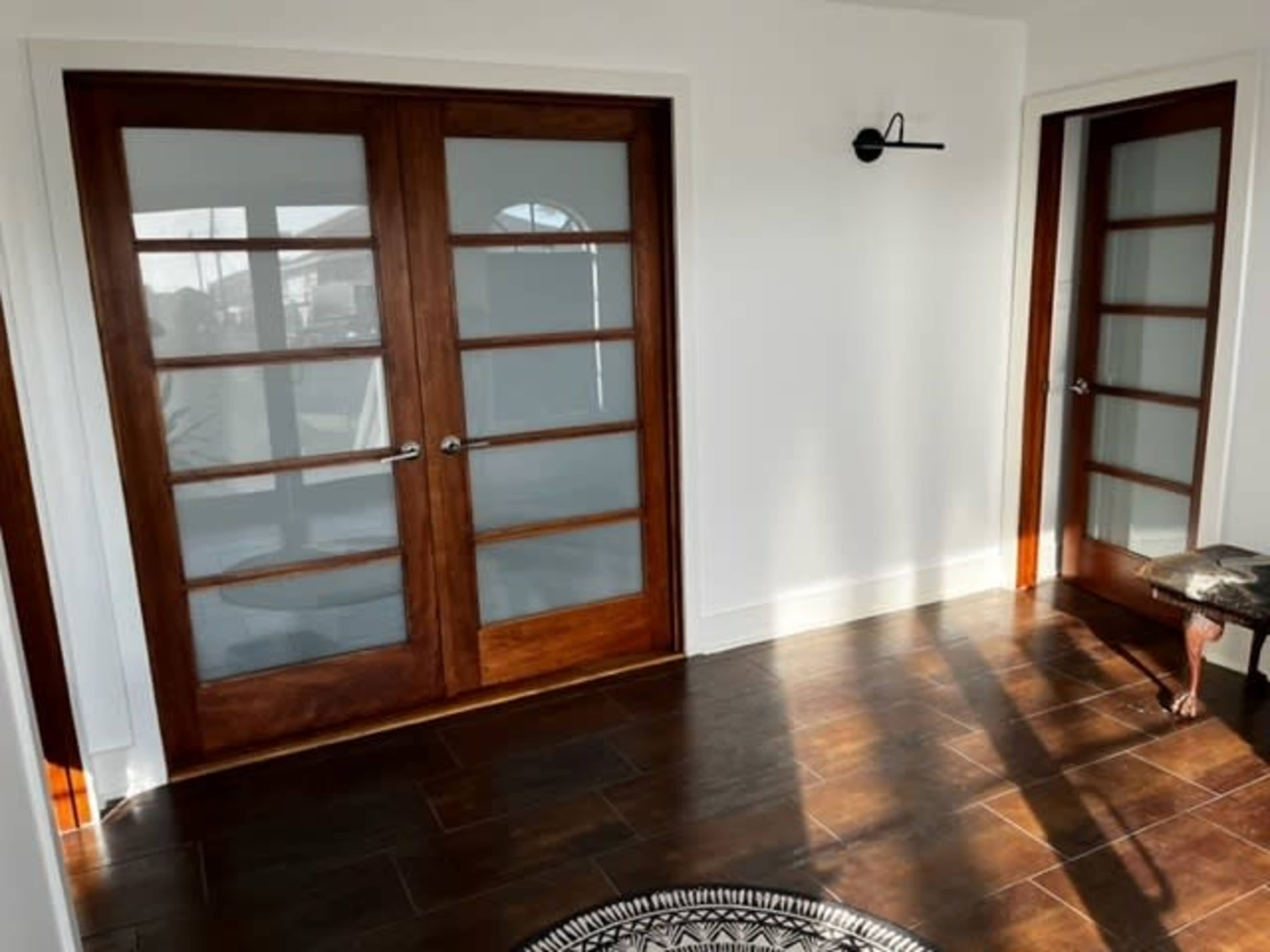 A brightly lit hallway featuring two wooden doors with glass panels, set against a backdrop of white walls and dark tiled flooring.
