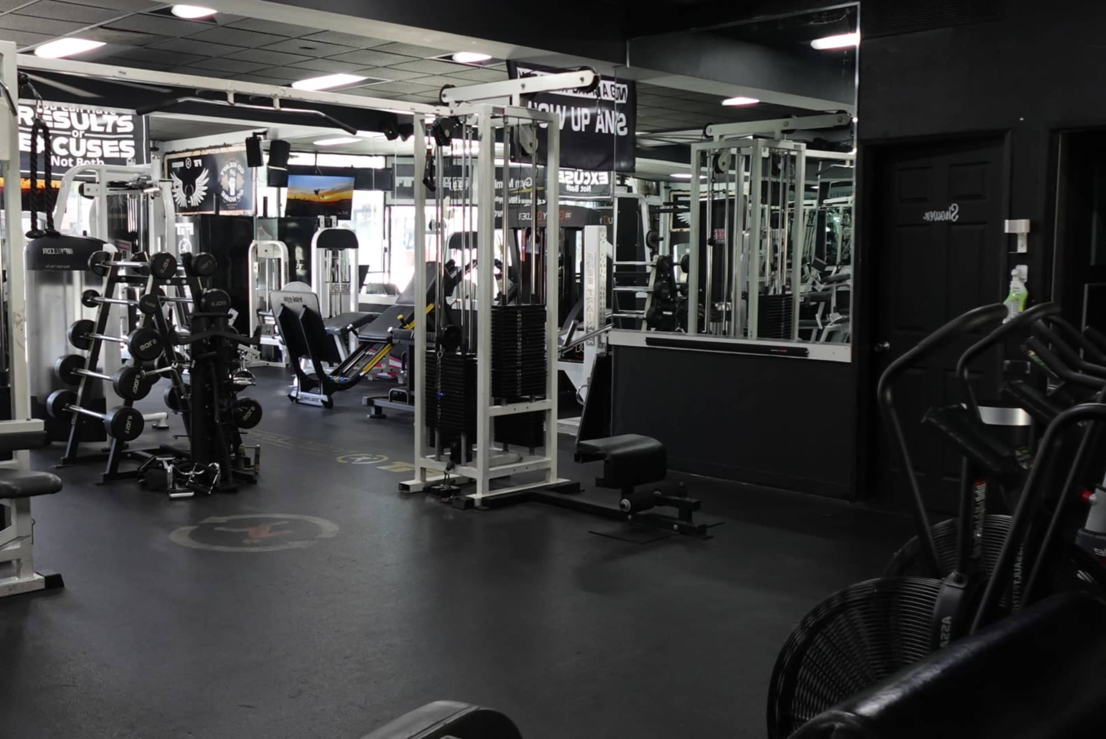 The image shows a gym interior with various workout equipment, including weight machines, benches, and cardio machines, arranged in a spacious layout.