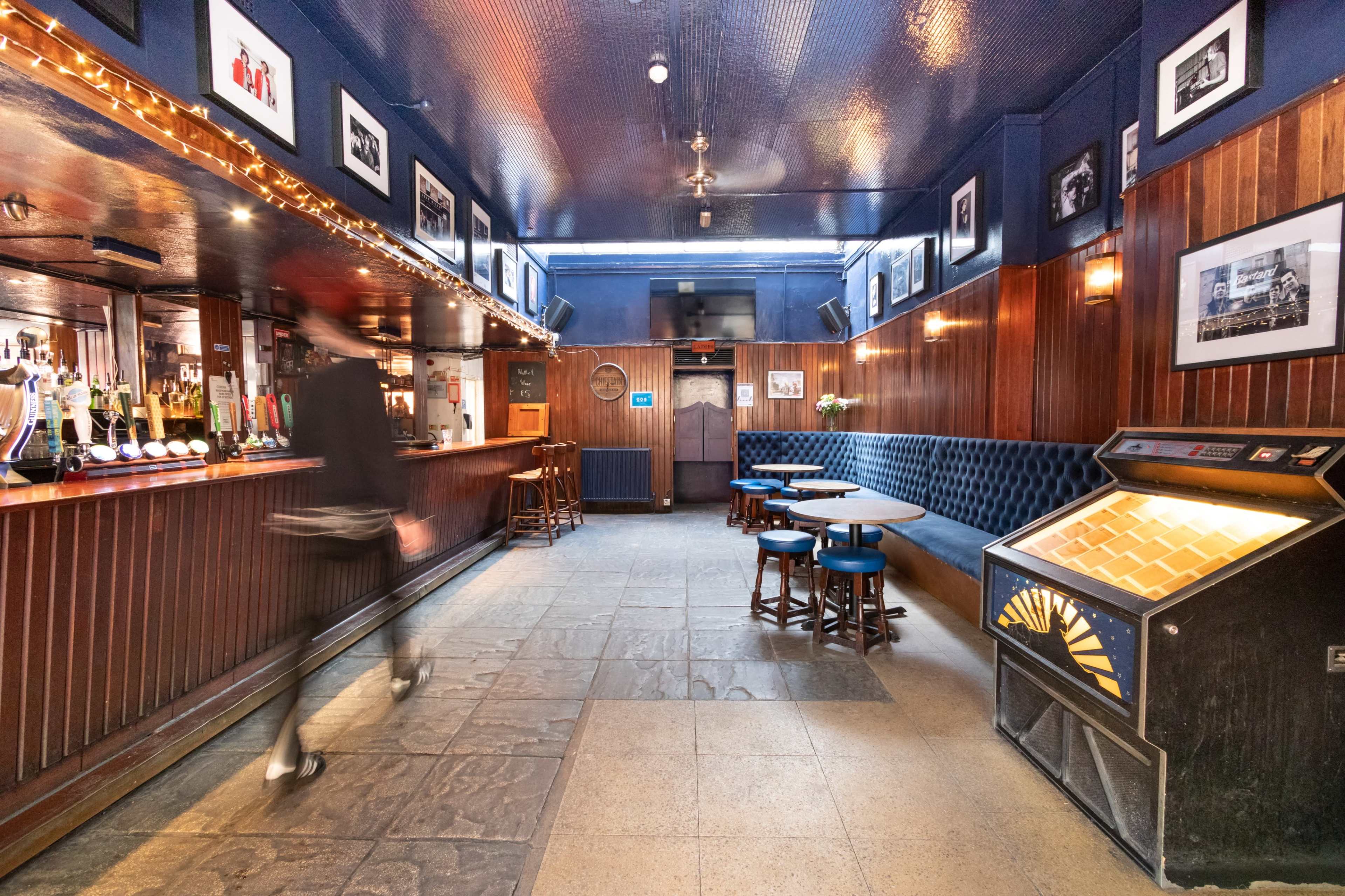 The image shows a dimly lit bar featuring a wooden counter, blue walls adorned with framed pictures, and a vintage jukebox in the corner.