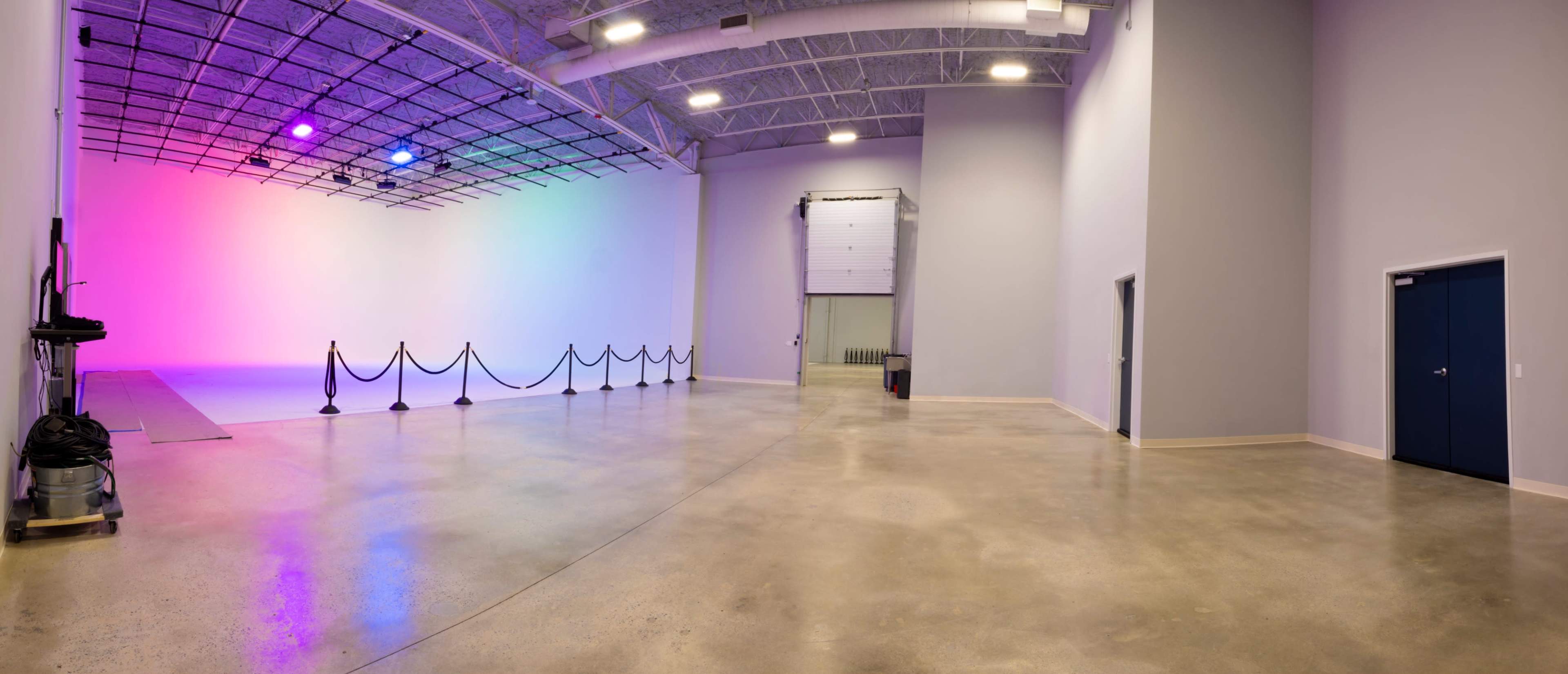 A spacious, well-lit studio with a gradient colored backdrop and a clean concrete floor, featuring ropes for crowd control and a large storage door in the background.