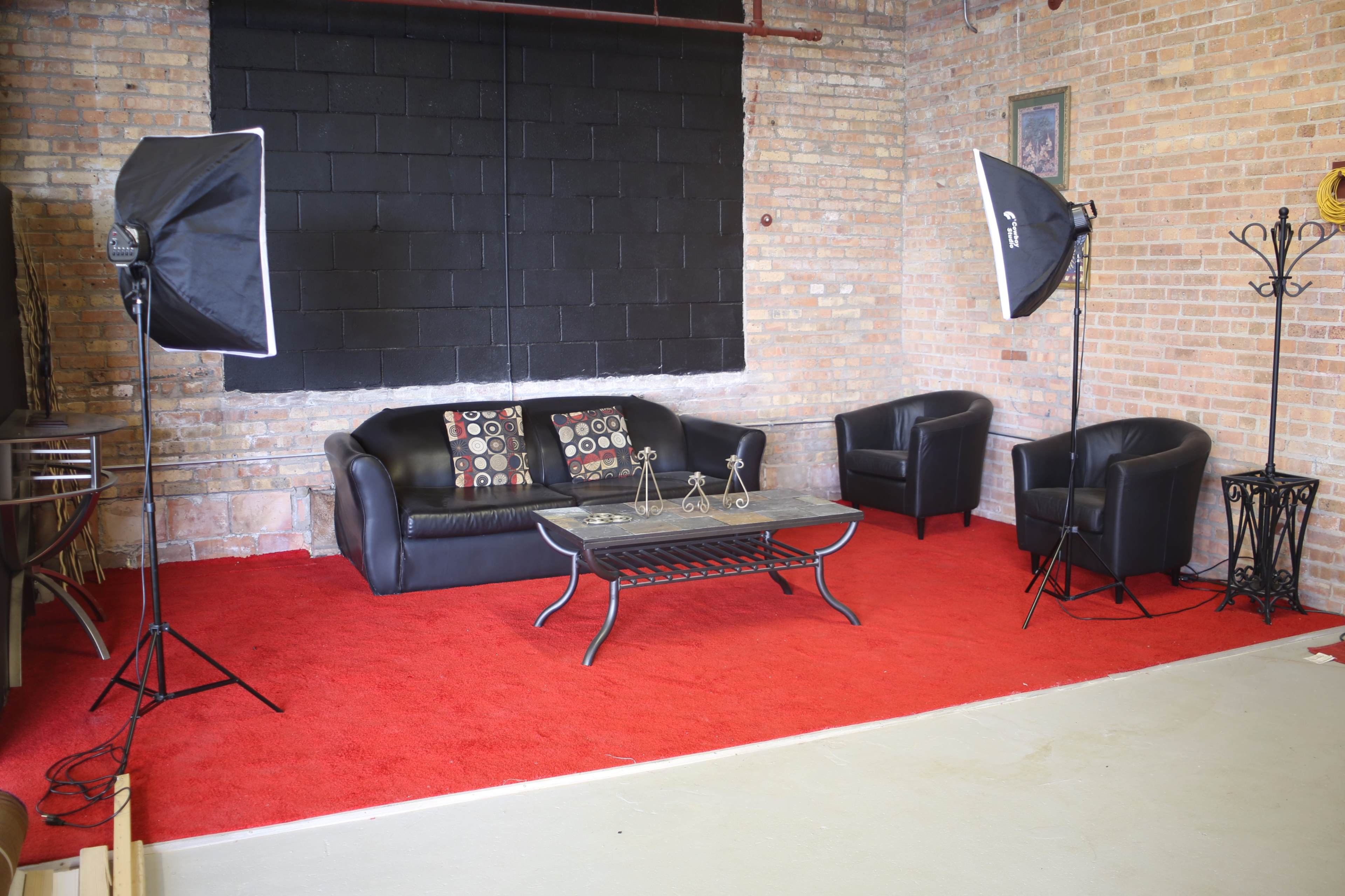 The image shows a seating area with a black sofa, two matching armchairs, a coffee table, and professional lighting equipment on a red carpeted floor against a brick wall.