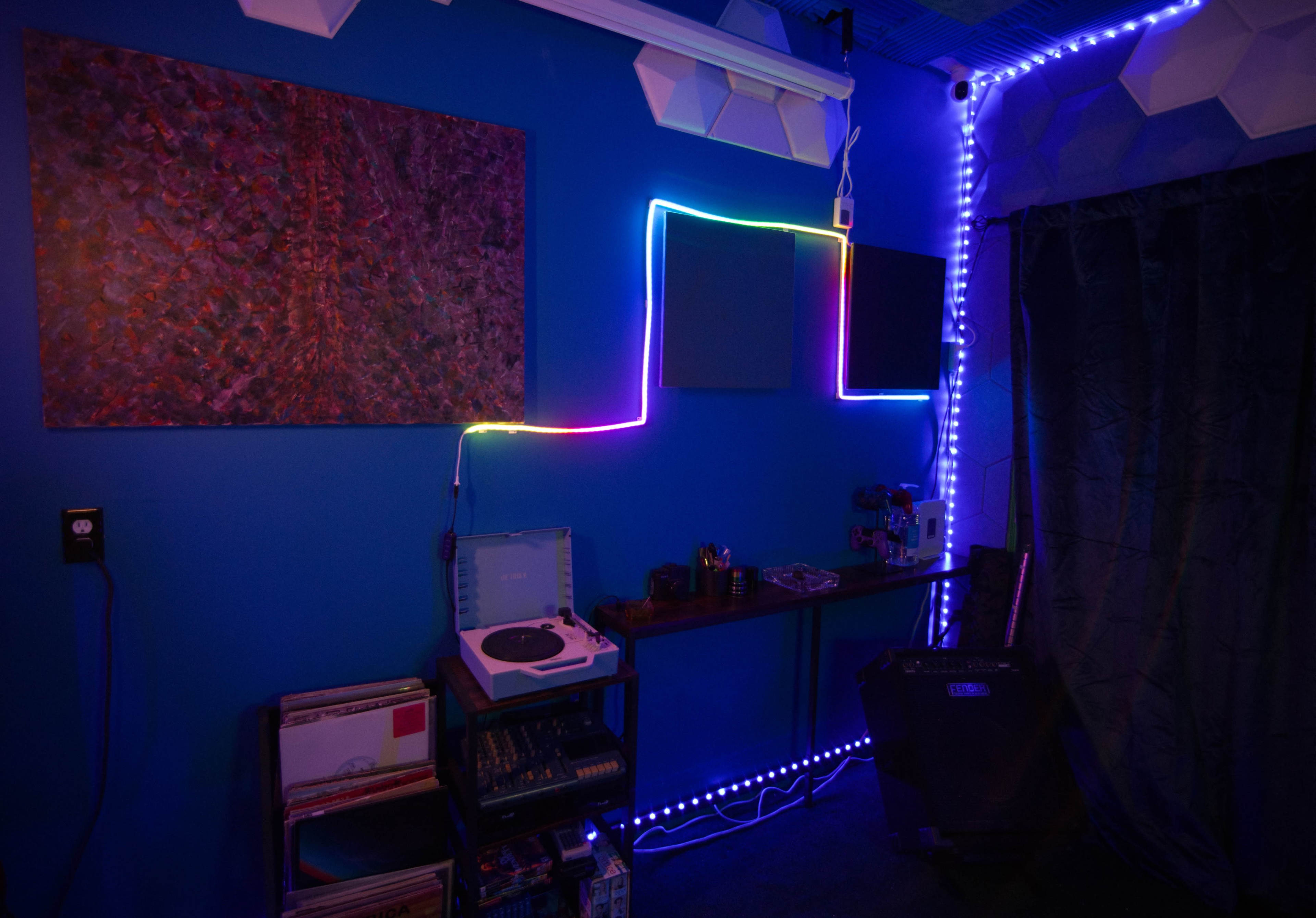 The image shows a room with a blue wall, a colorful neon light outline, a record player, and various electronic equipment on a shelf.