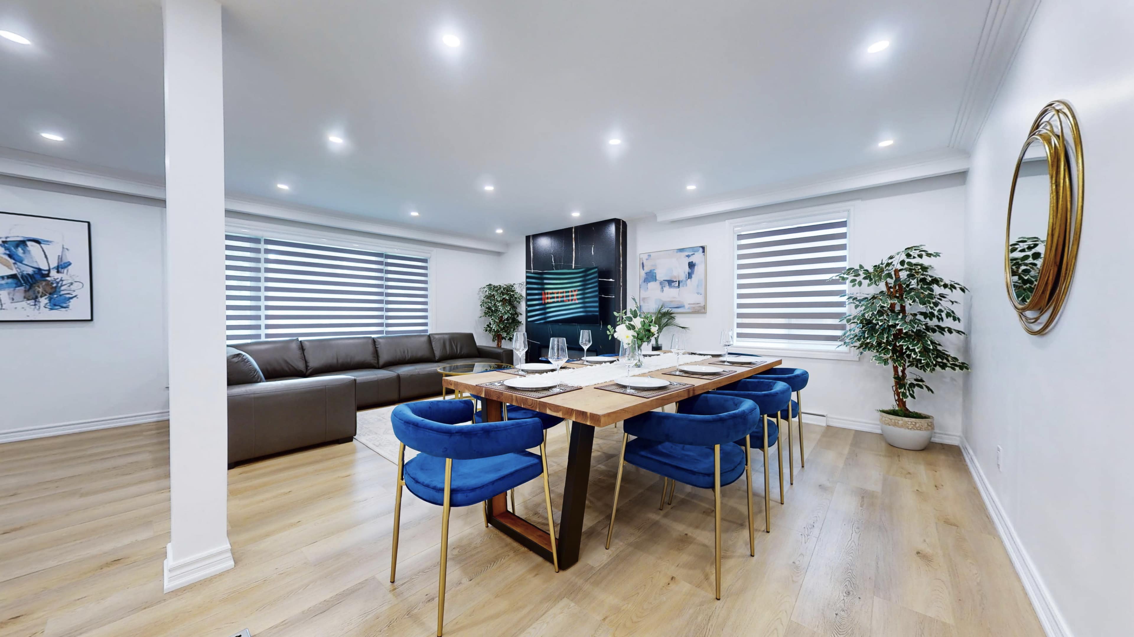 A modern dining area features a wooden table set for six, surrounded by blue chairs, with a sectional sofa and decorative plants in the background.