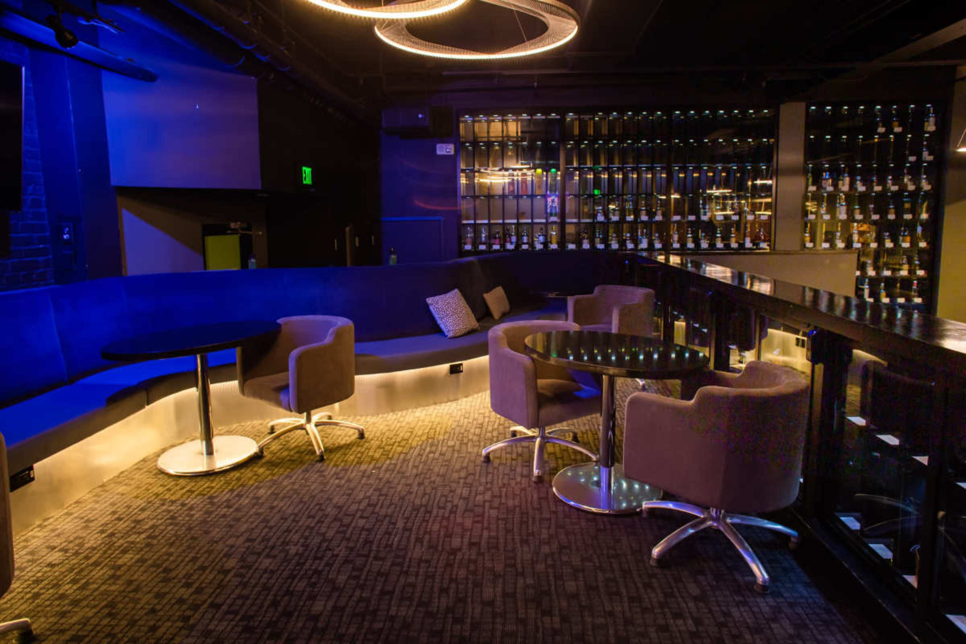 The image shows a dimly lit lounge with a curved seating area, circular tables, and a wall of bottles in the background.