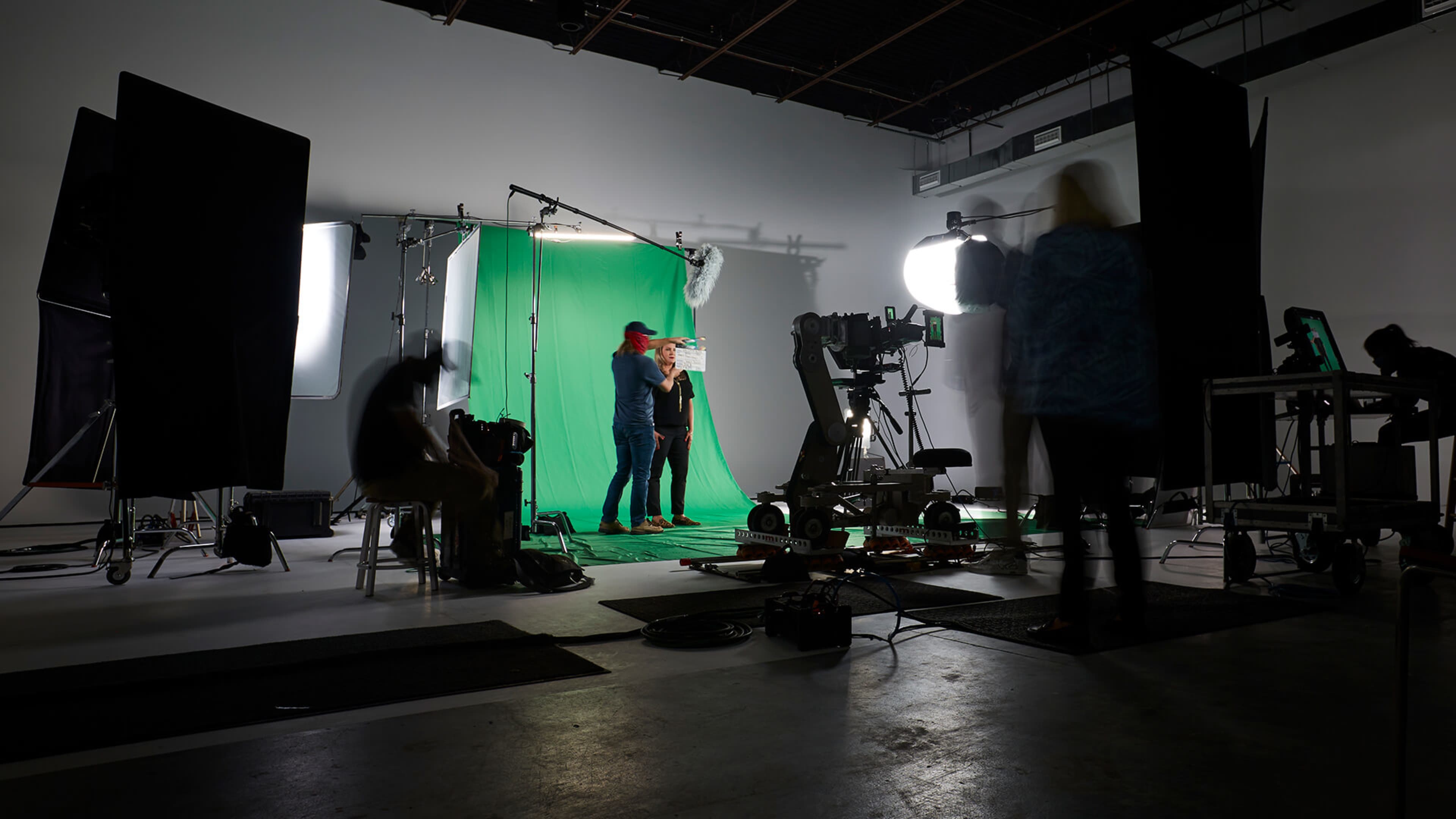 A film studio features a green screen setup with various lighting equipment and crew members preparing for a shoot.