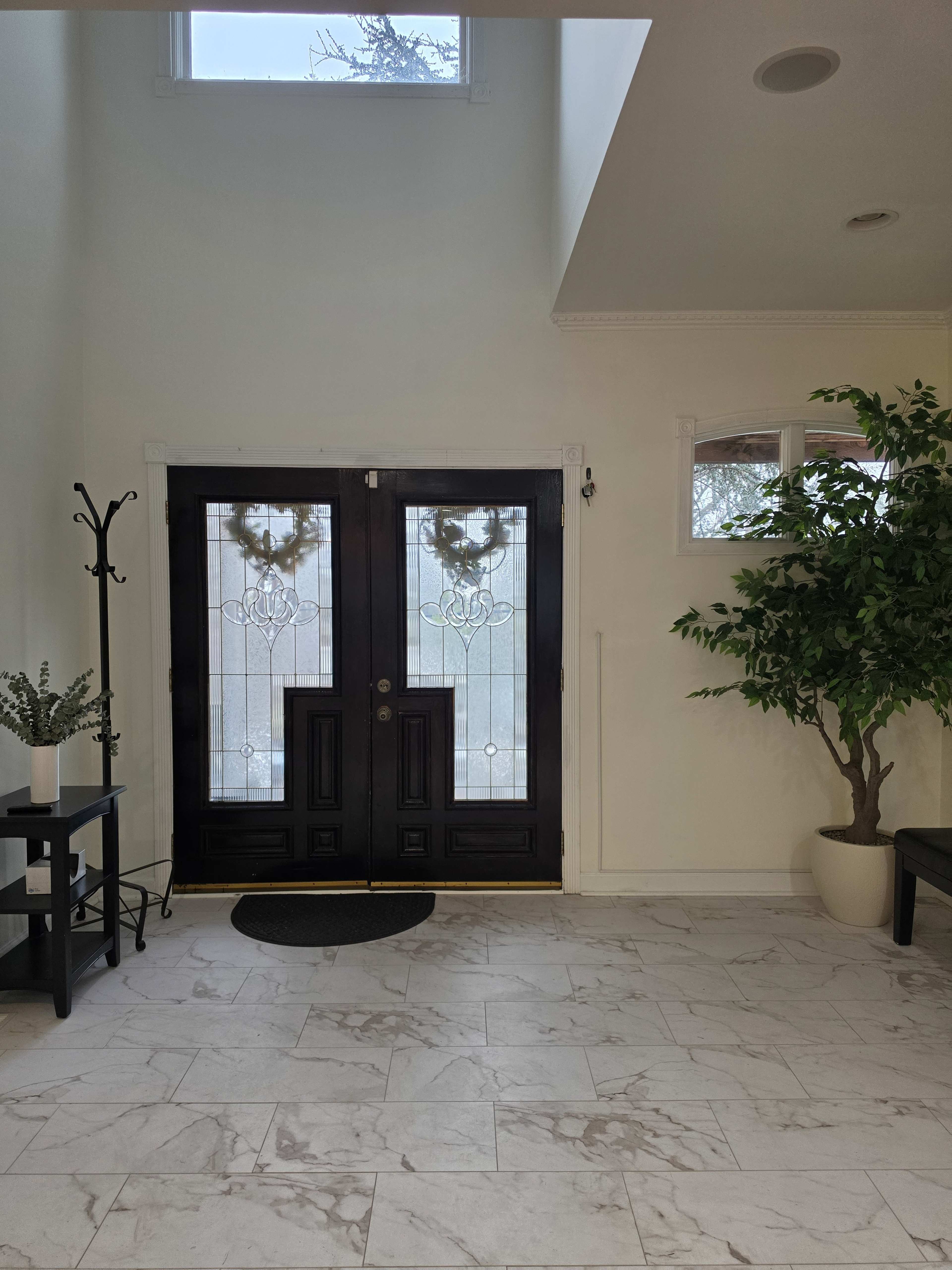 The image shows a spacious entrance hall featuring a pair of double doors with glass panels, a tall indoor plant in the corner, and marble flooring.