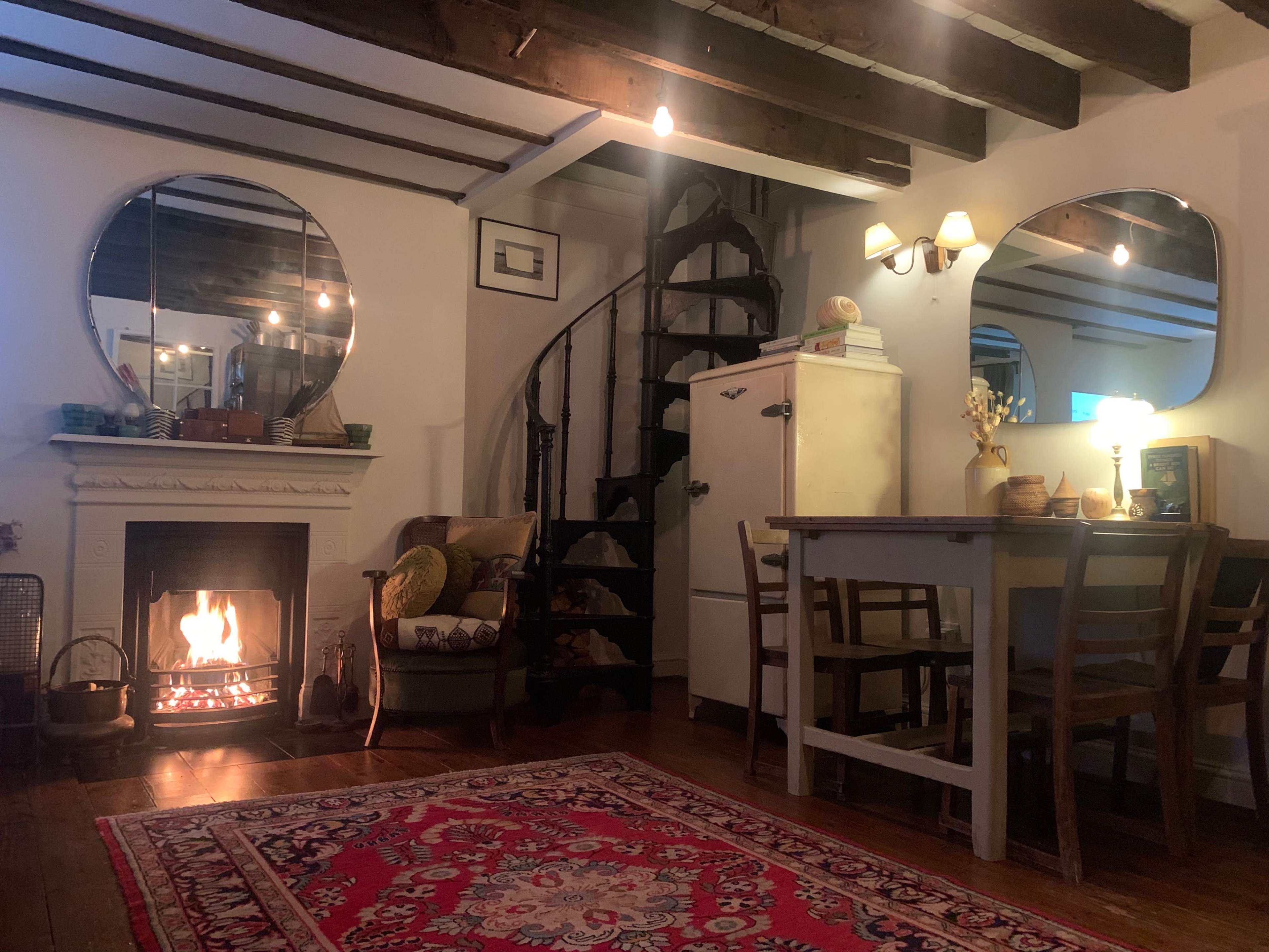 A cozy living space featuring a fireplace with a fire burning, a spiral staircase, two large mirrors on the walls, a dining table with chairs, and a sofa beside a rug on wooden flooring.
