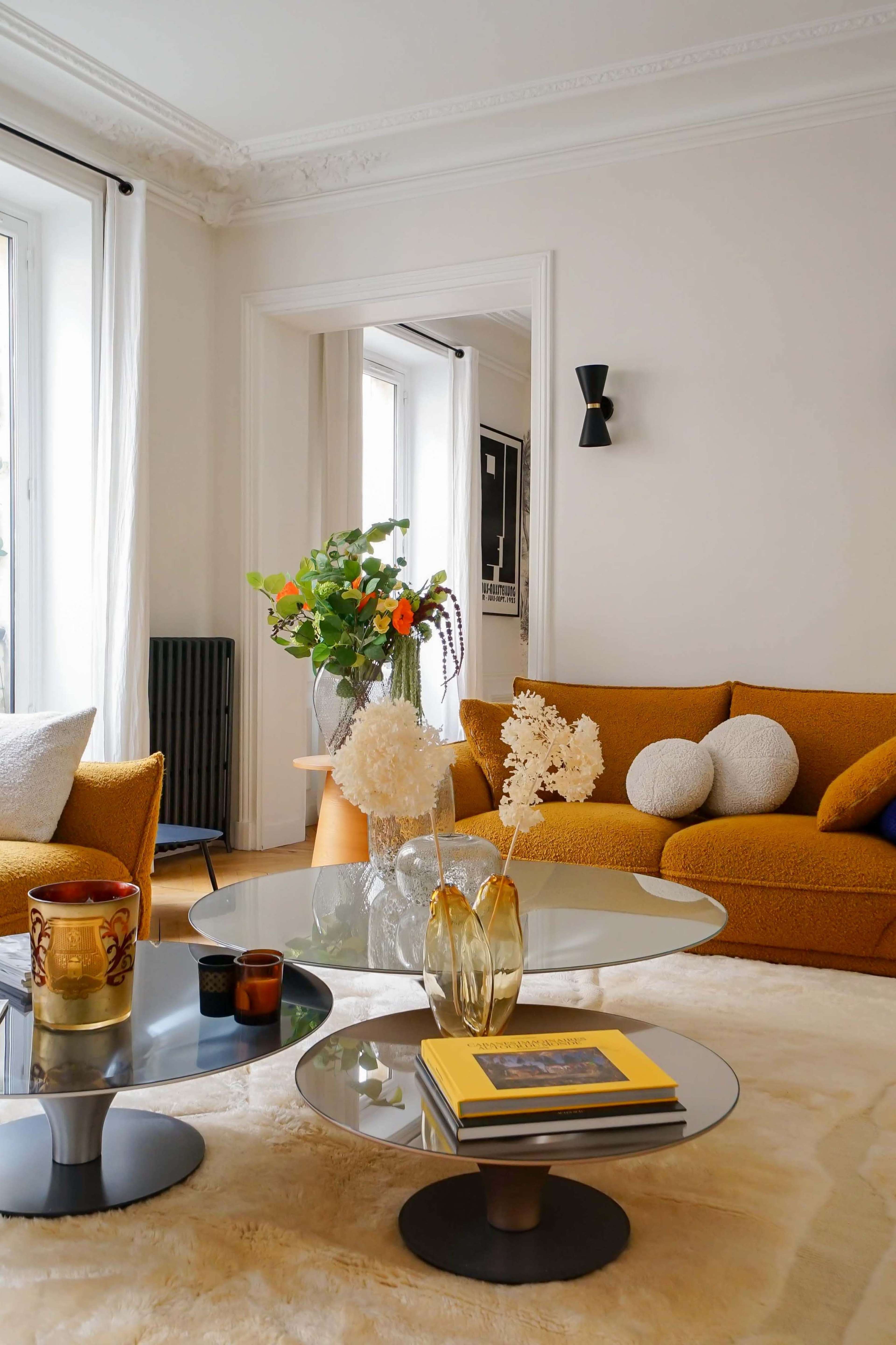 A cozy living room features a mustard yellow sofa, a glass coffee table with decorative items, and a vase of fresh flowers on a neutral-colored rug.