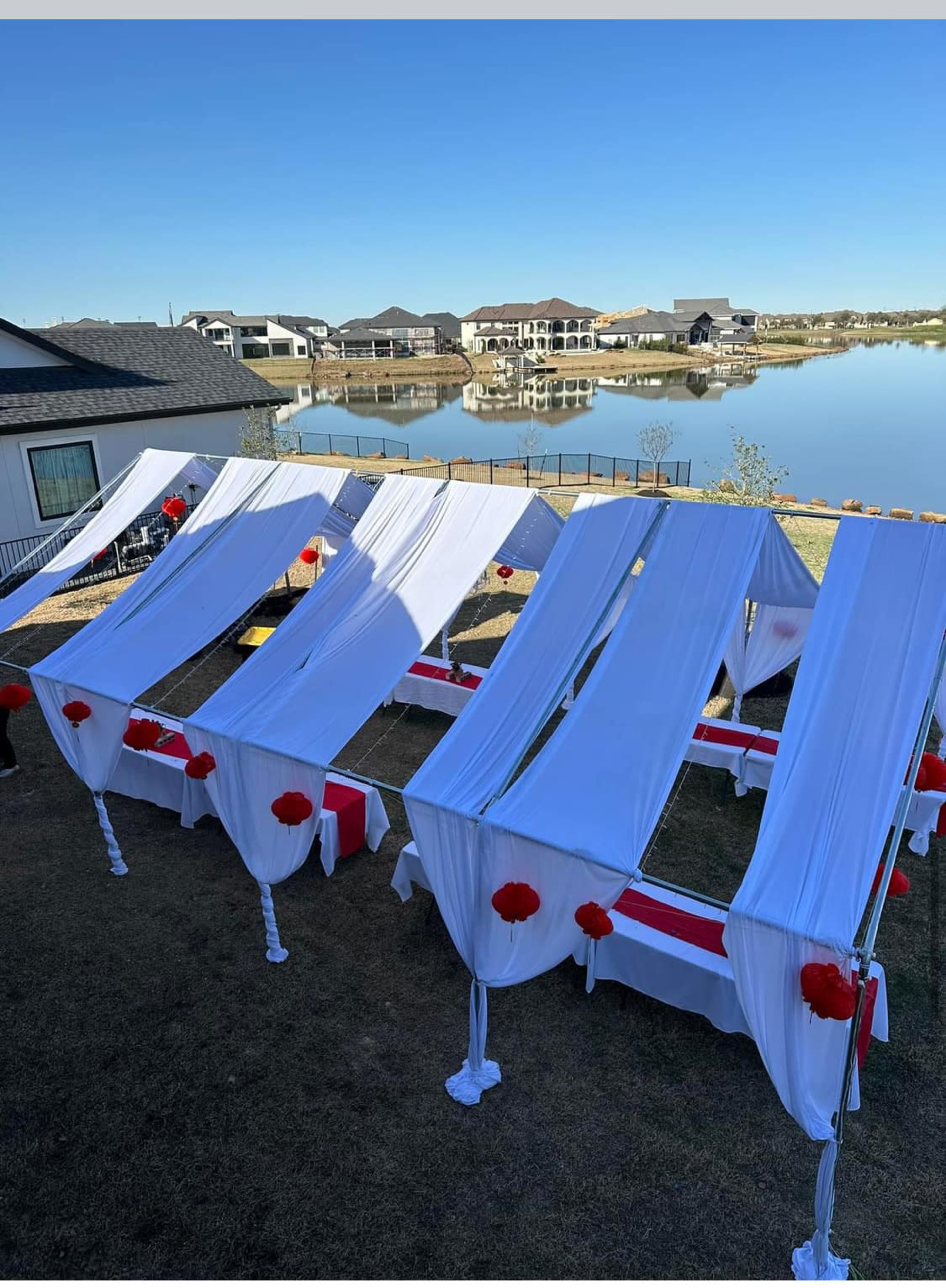 A setup for an outdoor event with several white canopies draped with red accents, positioned near a body of water and surrounded by houses in the background.