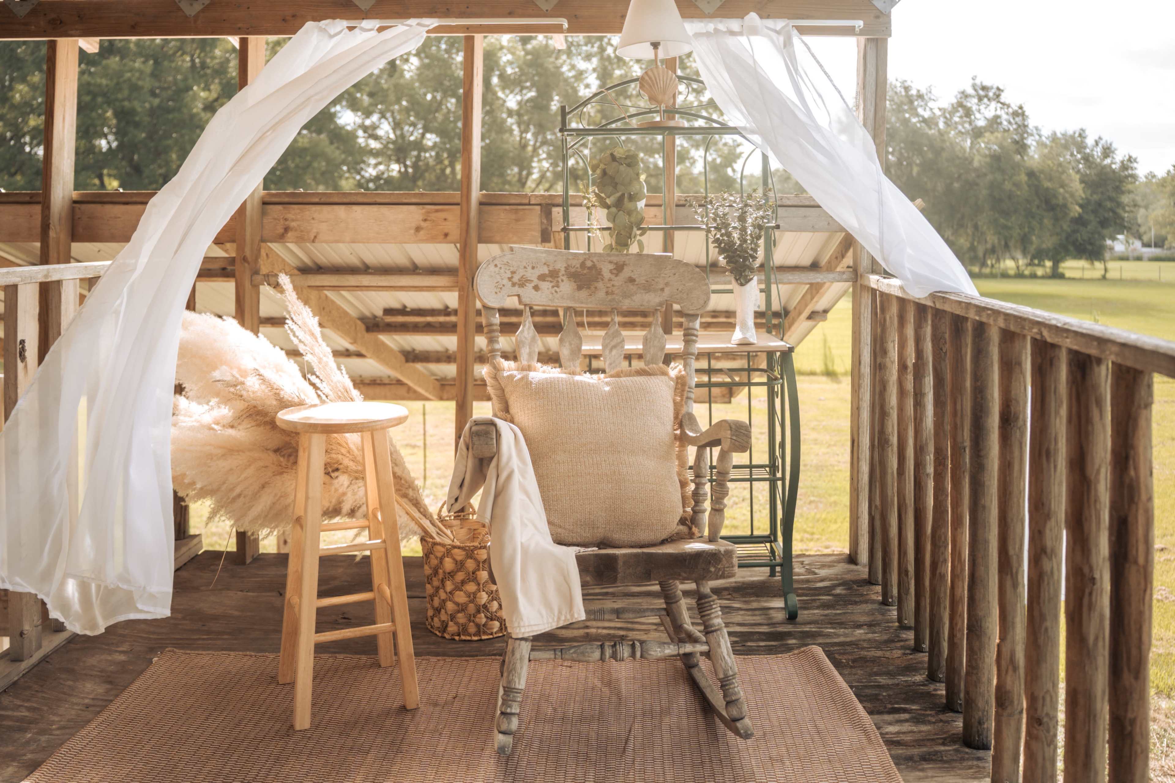 A wooden rocking chair sits on a deck beside a small stool, draped fabric billows in the breeze against a backdrop of trees.