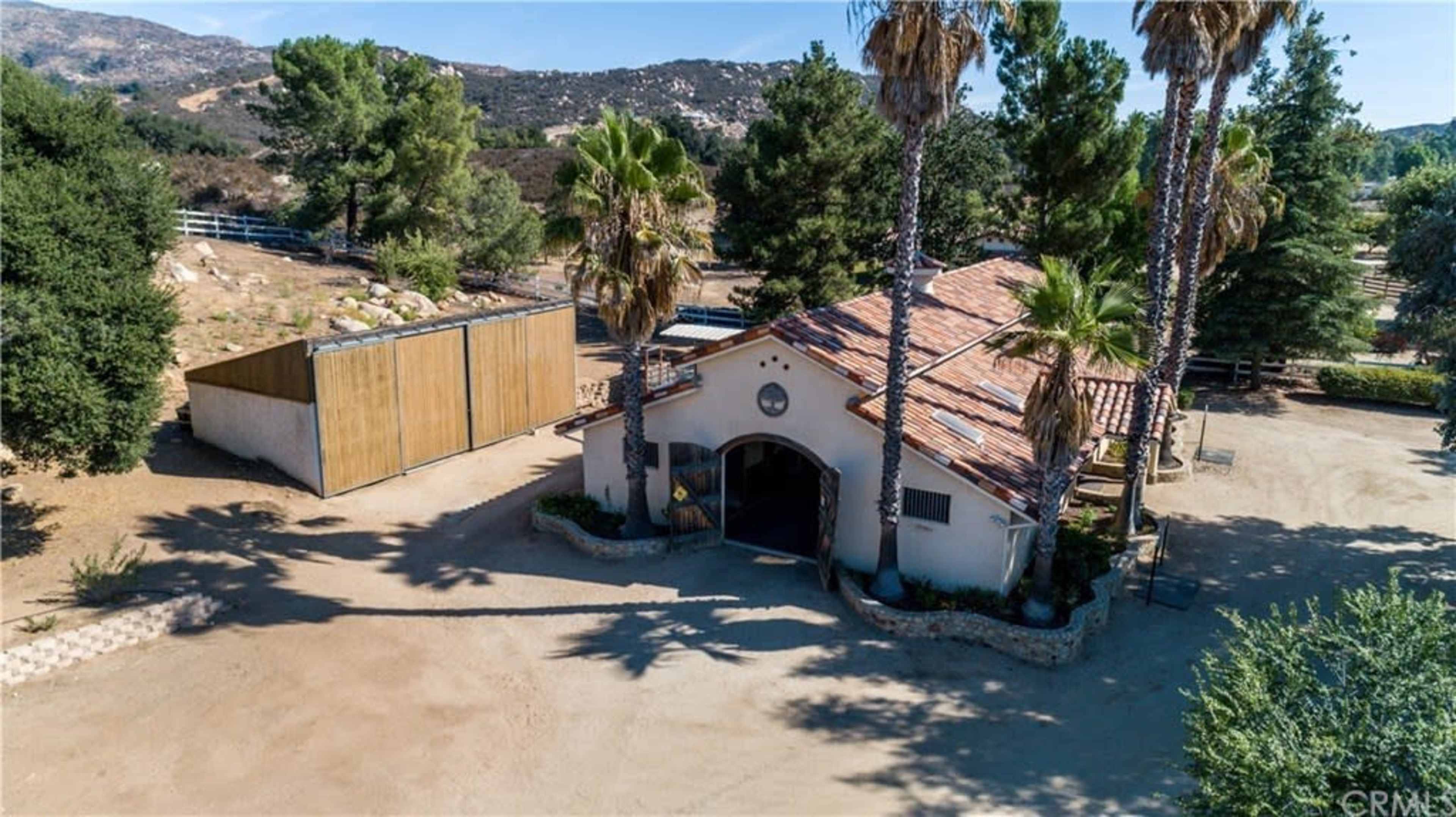The image shows a single-story building with a red-tiled roof surrounded by palm trees and dirt paths, set against a mountainous landscape.
