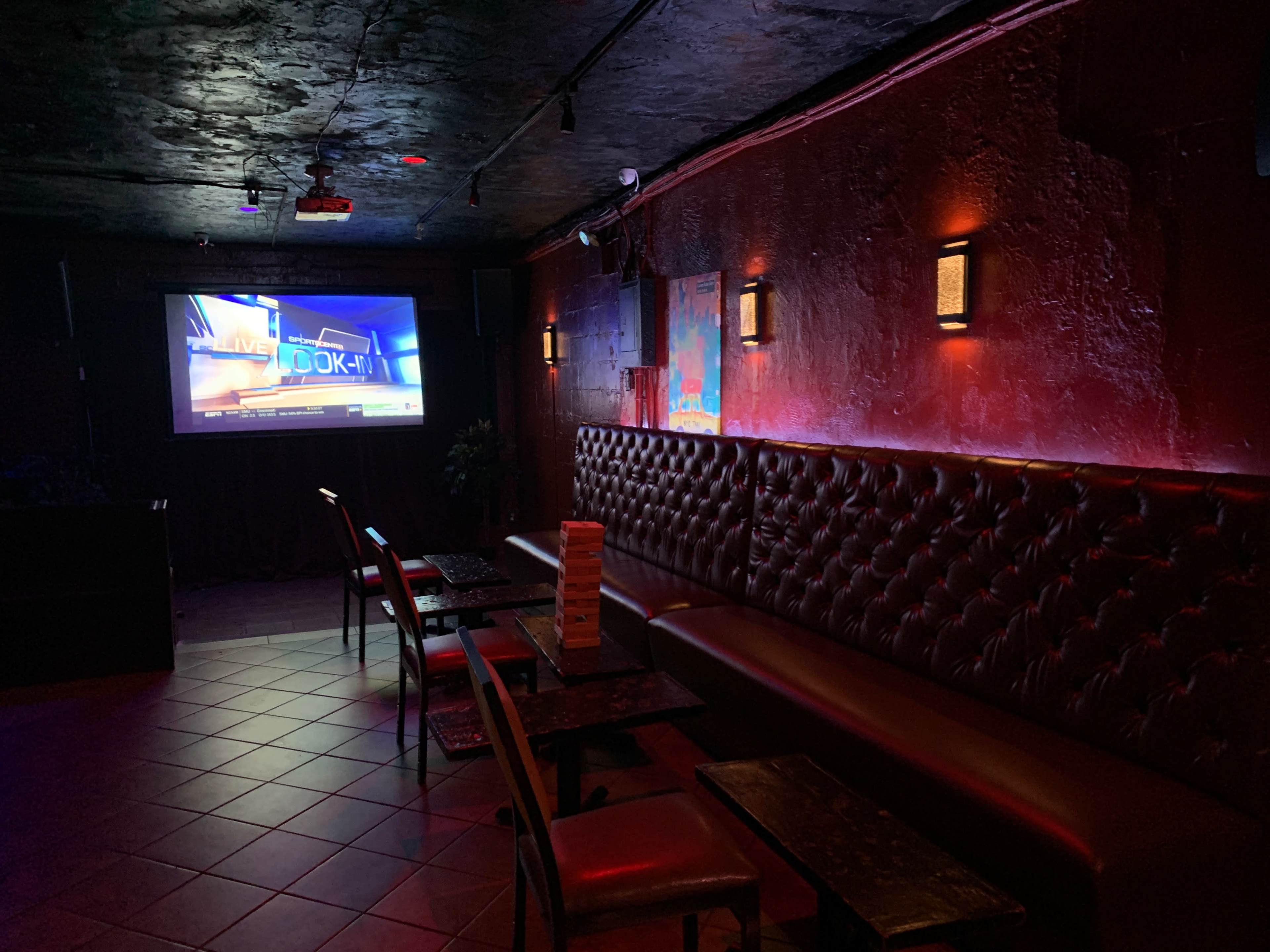 A dimly lit lounge area with a large screen displaying a sports broadcast, featuring leather seating and small tables.