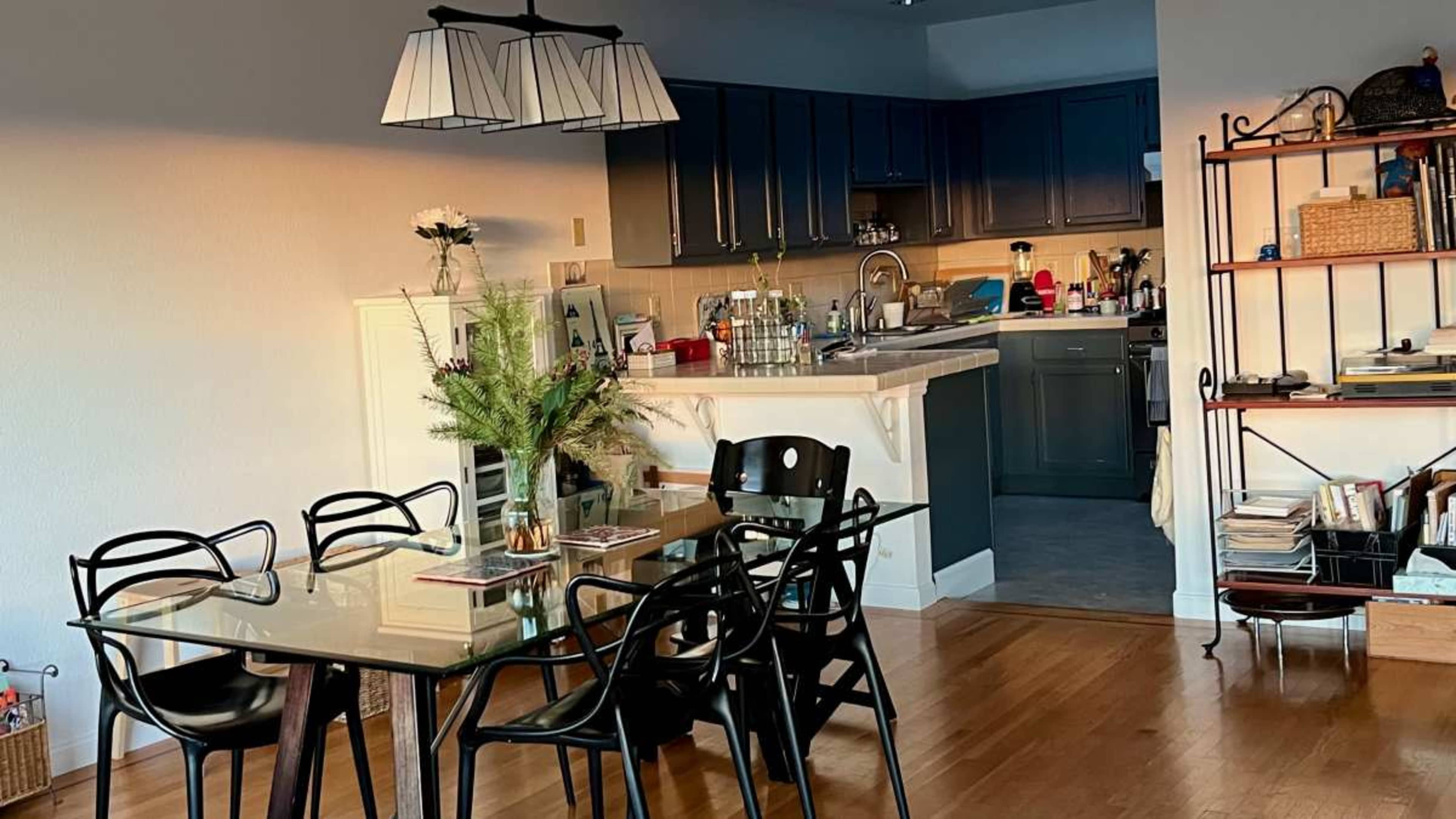 The image shows a dining area with a glass table and black chairs, adjacent to a kitchen featuring dark cabinetry and various kitchen items visible.