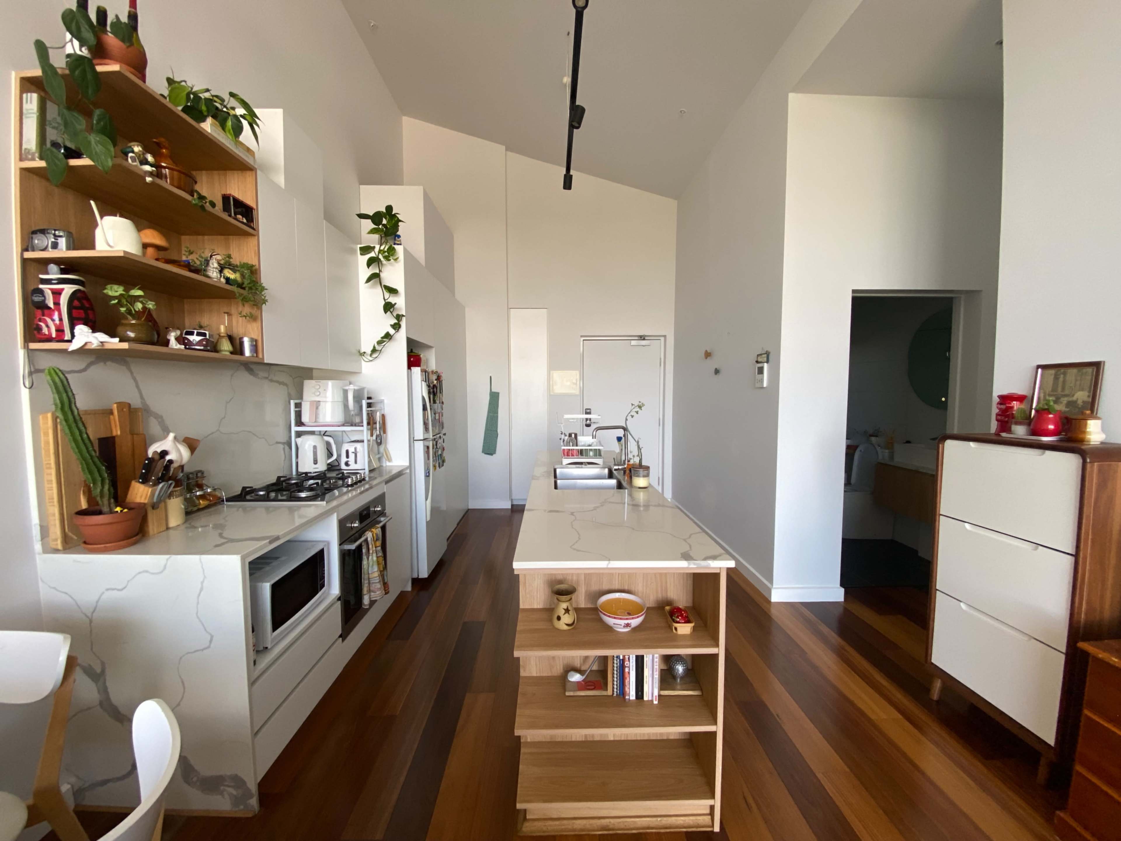 The image shows a modern kitchen with a central island, open shelving displaying plants and kitchenware, and a mix of wooden and white cabinetry.