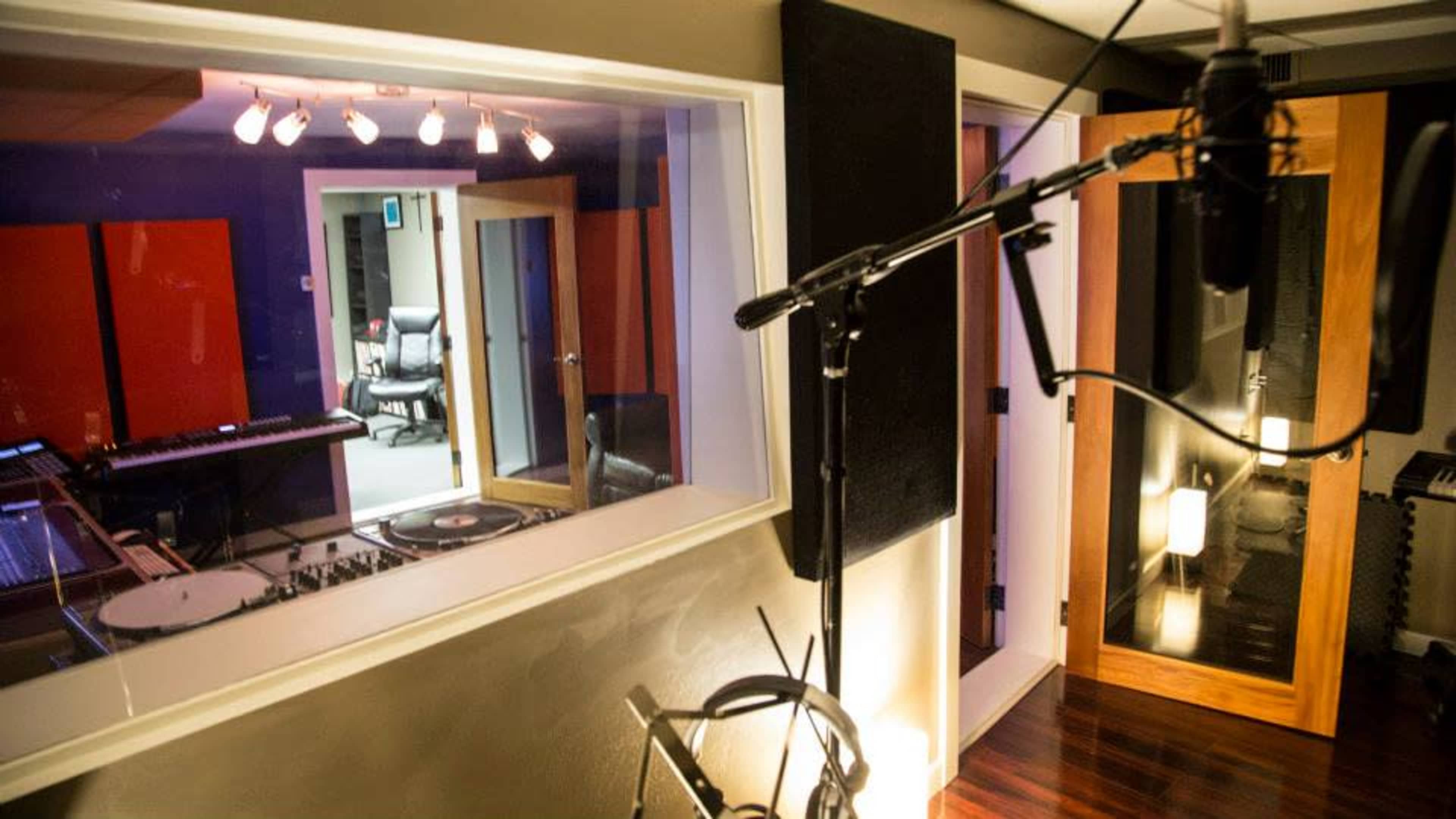 A sound recording studio featuring a microphone in the foreground and a control room visible through a glass wall.