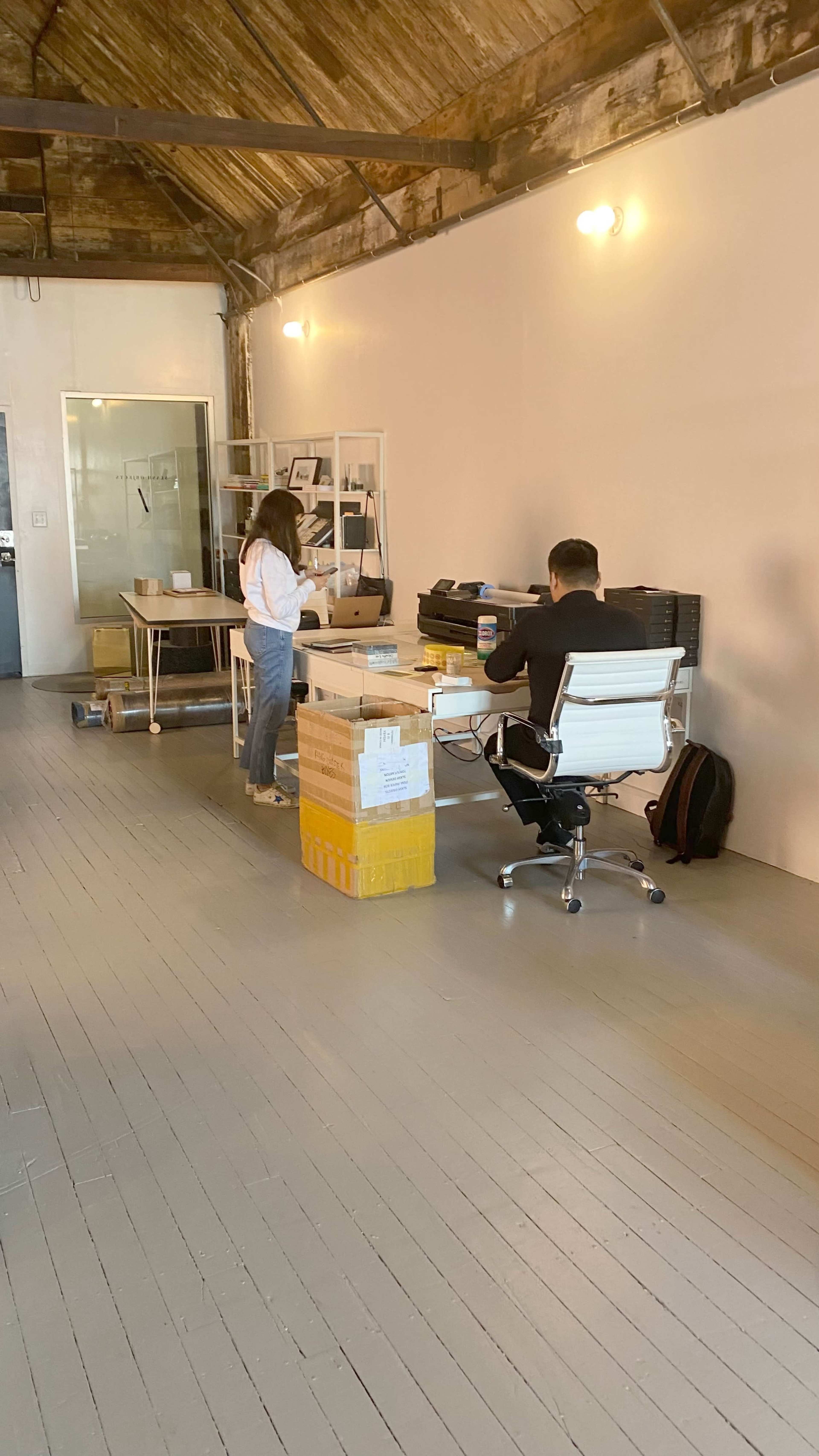 Two individuals work in a modern office space with a large table, storage boxes, and minimalist furniture.