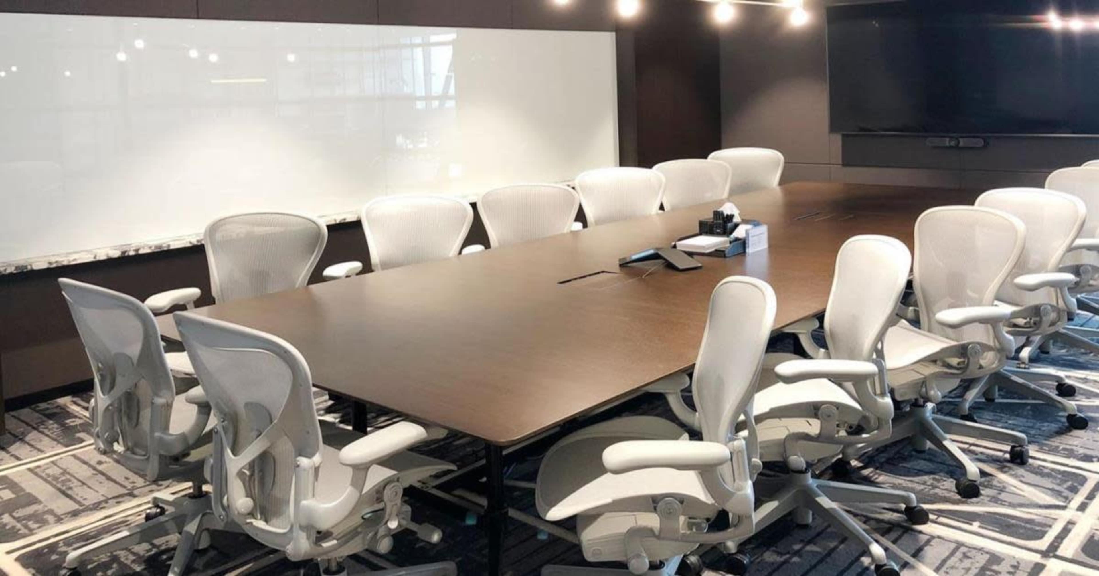 A large conference room features a rectangular table surrounded by multiple ergonomic chairs, with a whiteboard and a screen on the walls.