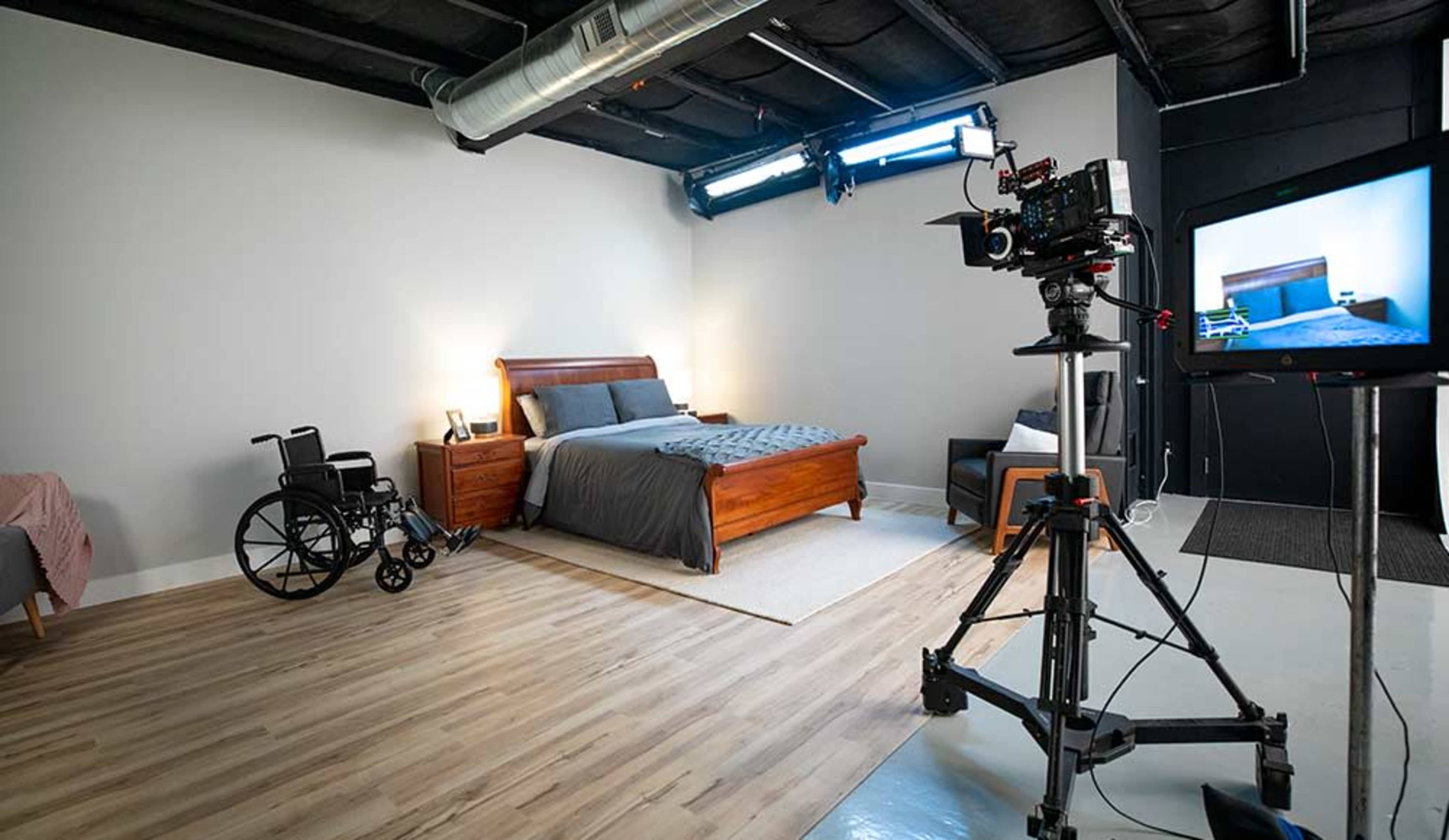 A well-lit bedroom setup features a wooden bed, a wheelchair, and a camera on a tripod with a monitor displaying the scene.