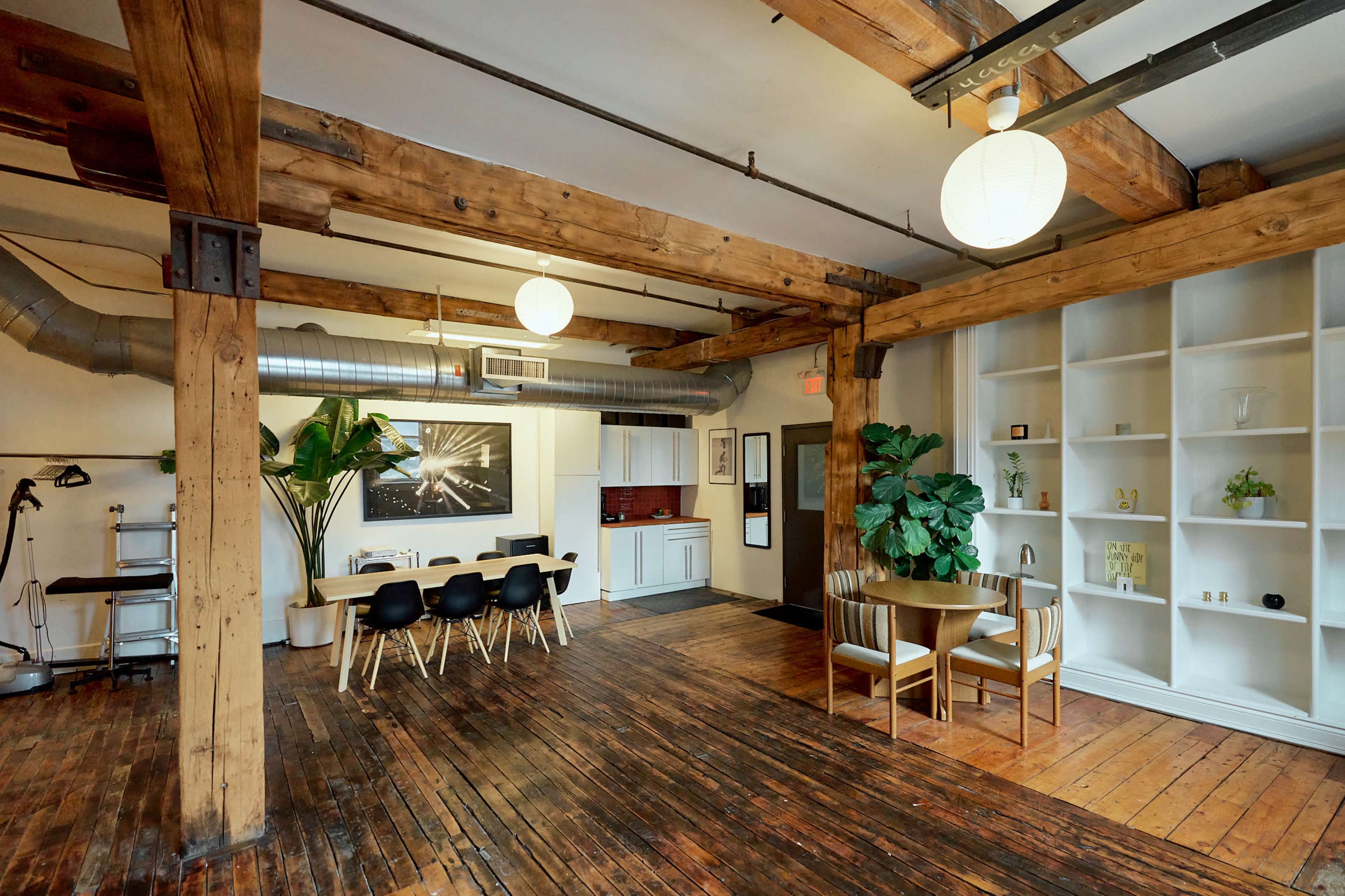 The image shows a bright, open room with wooden beams and floors, featuring a dining area with a table and chairs, a kitchen space, and a decorative plant in the corner.