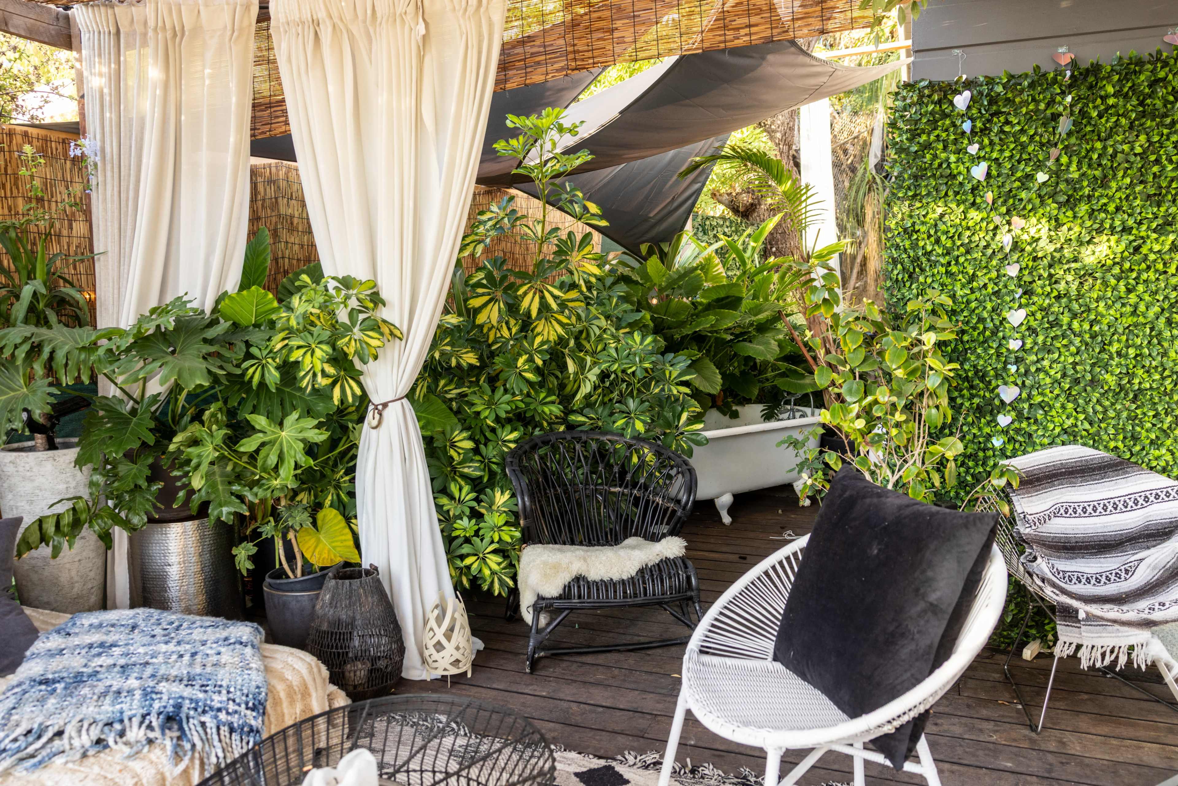 The image shows a cozy outdoor space with various potted plants, white curtains, and a mix of furniture, including a bathtub and lounge chairs.