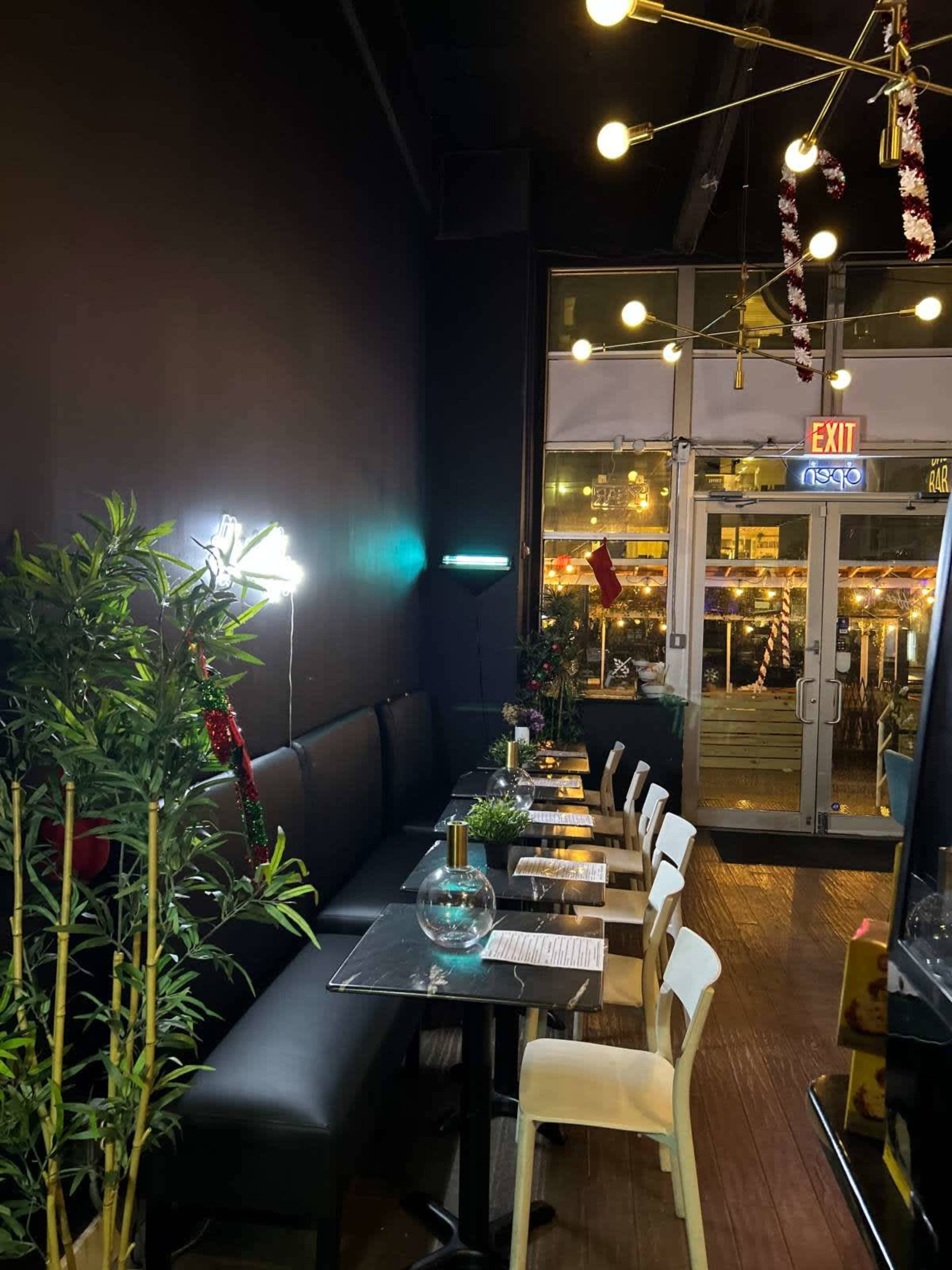A dimly lit restaurant interior with black walls, a long table set against a window, and decorative plants alongside white and black chairs.