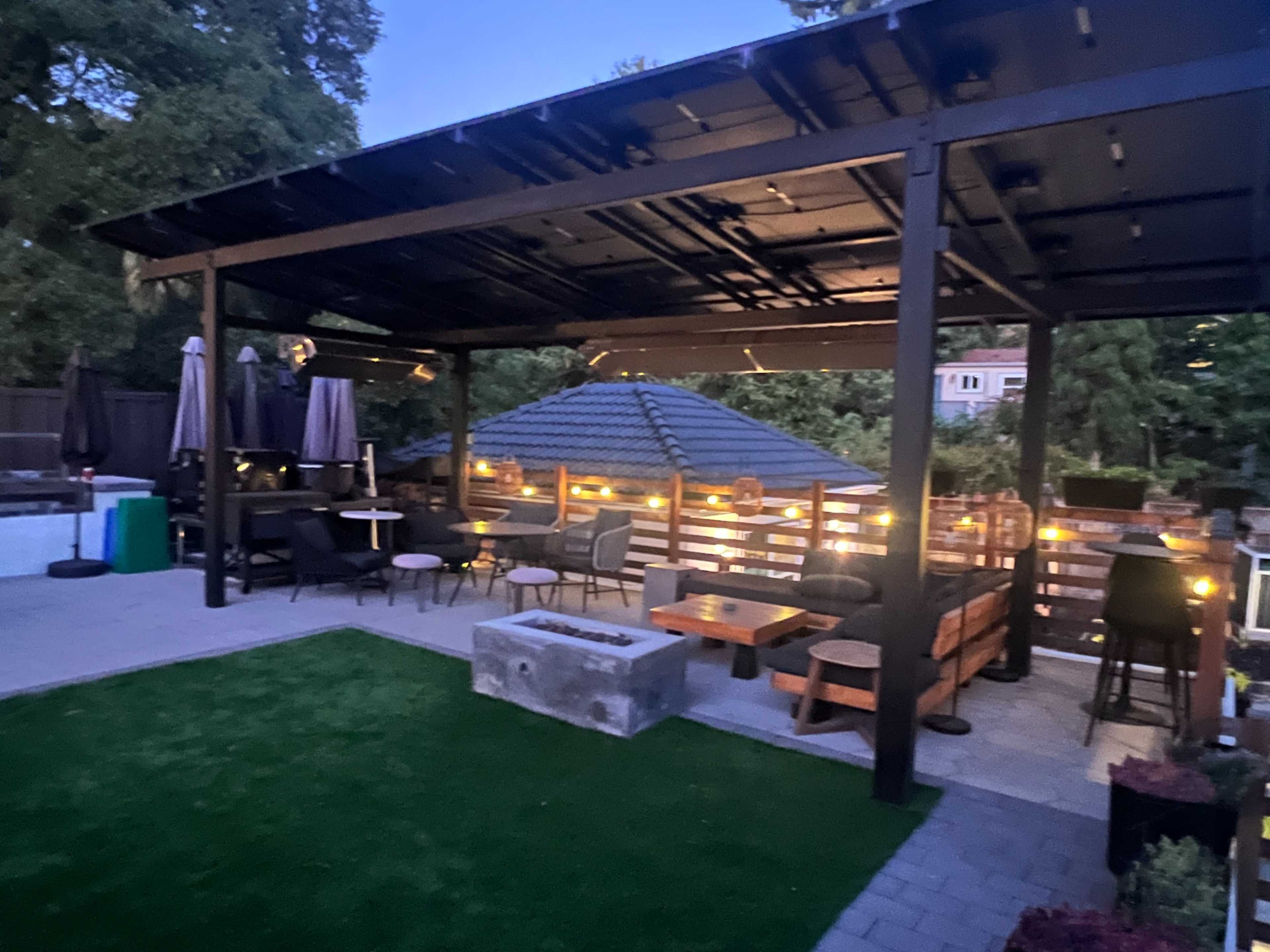 The image shows an outdoor patio area with a covered seating space, surrounded by greenery and soft lighting.